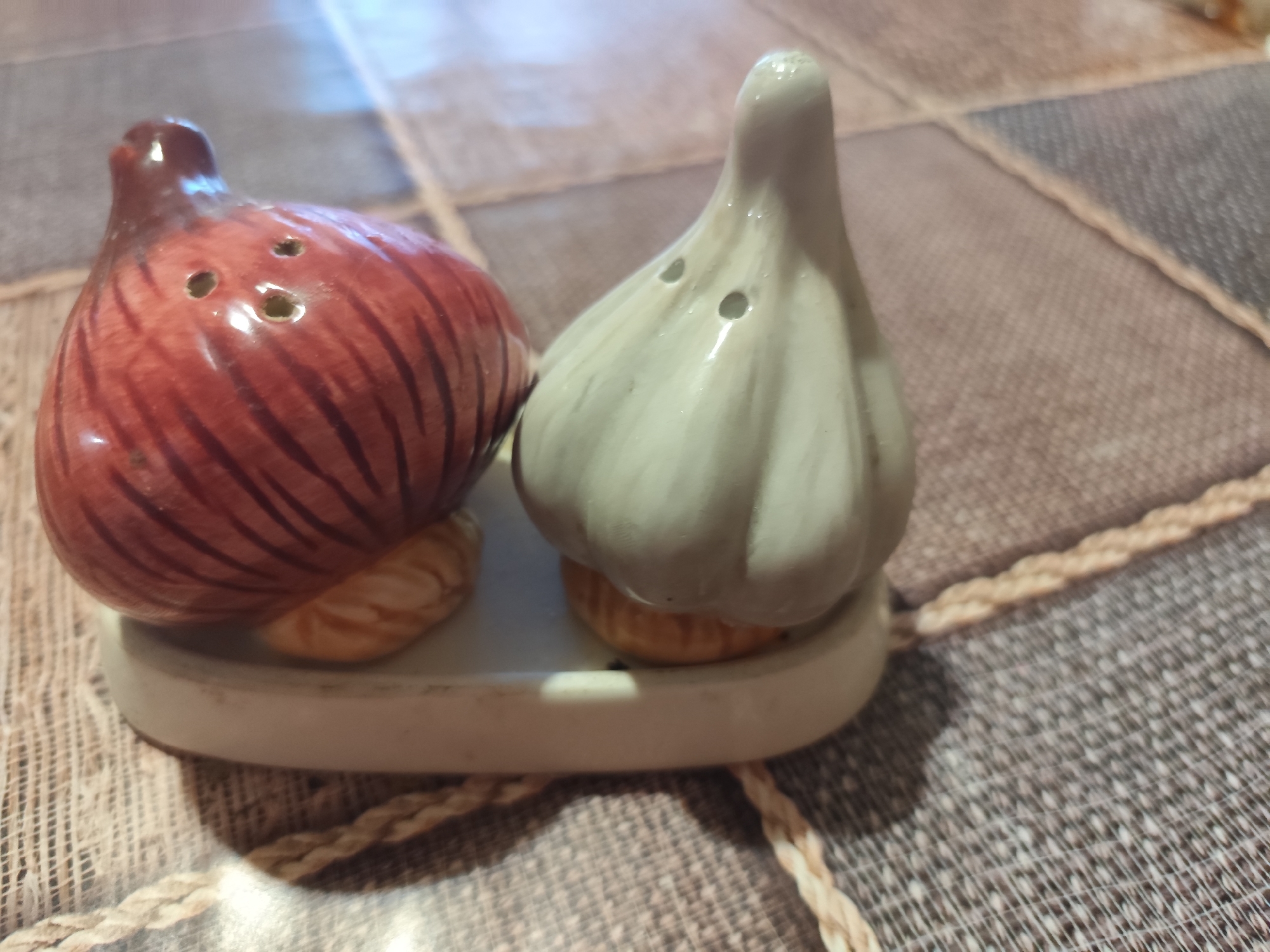 An overhead shot shows a pair of salt and pepper shakers, shaped like an onion and a garlic bulb, resting on a white ceramic tray. The shakers are ceramic, with the onion shaker being red with dark brown stripes and the garlic shaker being light green with subtle vertical ridges. Both shakers have small holes at the top for dispensing spices. The tray sits on a woven placemat with a patterned design. The background shows a tiled surface, possibly a tabletop. There are no people or animals present in the image. The scene appears to be set indoors. The location of Montana, Bulgaria is not determinable from the image.