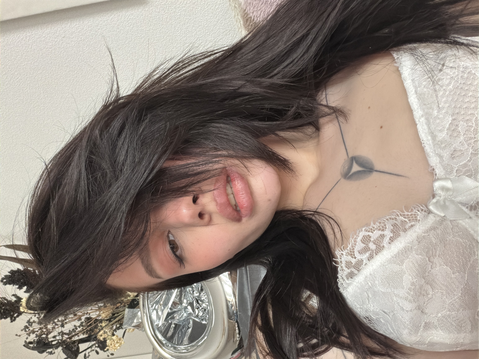 A close-up, angled portrait shows a woman with long, dark hair. She is wearing a white lace top. A tattoo is visible on her chest. The background includes dried flowers and a reflective object. The location is Unknown, Unknown.