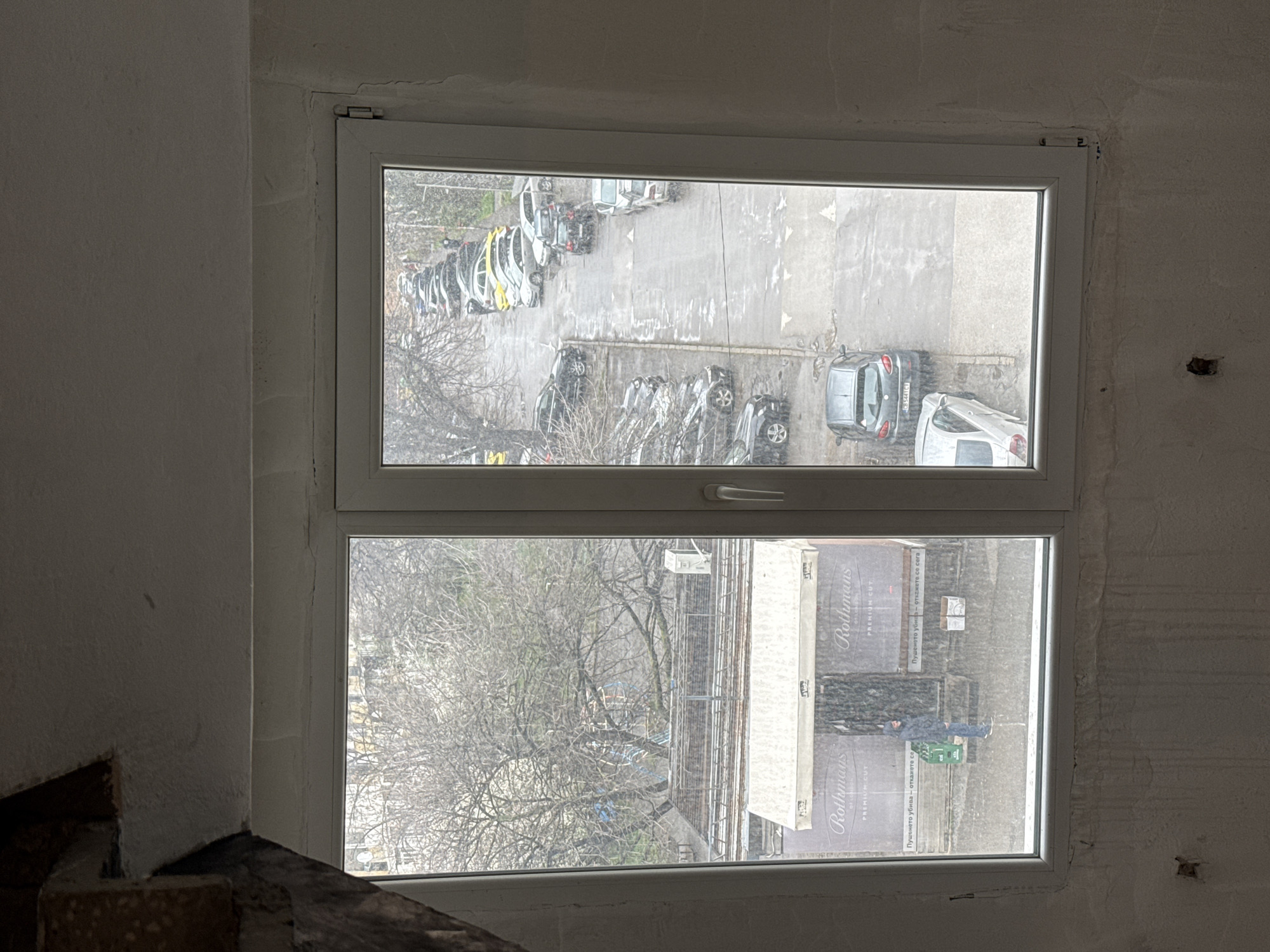 The image shows a view from a window in what appears to be an unfinished or partially renovated room. The window is a double-paned unit with a white frame.
Outside, the scene suggests an urban environment, possibly Sofia, Bulgaria, based on the Cyrillic lettering visible on an advertising banner. The top window offers a view of a parking lot filled with numerous cars. The lower window reveals a tree with bare branches, a building, and an advertising banner with the brand "Rothmans" and Cyrillic text. A person wearing blue jeans and a jacket, with what appears to be a green object next to them, is visible in front of the banner.
The room itself has rough plaster on the walls and exposed brick or concrete in the lower left corner of the image. The overall impression is one of a building undergoing construction or renovation.