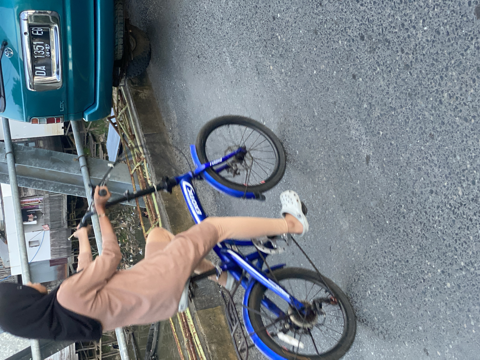 A person wearing a black hijab and a light brown outfit is riding a blue bicycle on a paved road. The bicycle has a "EXOTIC" logo on the frame. The rider's left foot is on the pedal, and they are wearing white clogs. A teal SUV with a license plate that reads "DA 1351 EG" is parked to the right of the road, partially visible. In the background, there are buildings on stilts over a body of water. The scene appears to be located in Tunggul Irang Ulu, Indonesia, based on the license plate format which is common in Indonesia. The road surface is asphalt with visible aggregate. The lighting suggests it is either early morning or late afternoon.