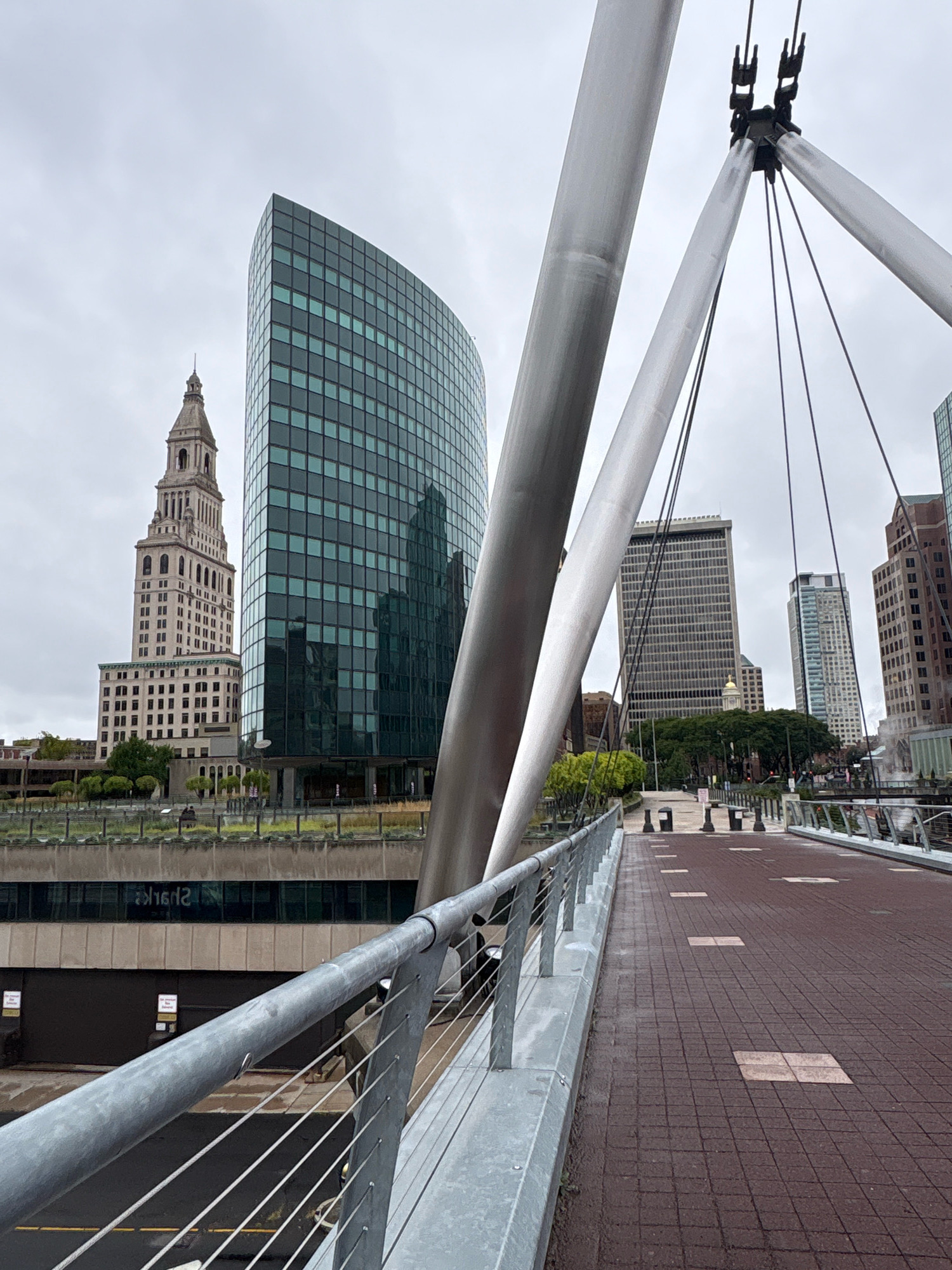 The image shows a view of the skyline of Hartford, Connecticut, from a pedestrian bridge. In the foreground, the sleek metal cables and supports of the bridge are visible. The bridge deck is paved with reddish-brown brick pavers, and a metal railing runs along the side.
In the background, several buildings rise above the cityscape. The most prominent is a tall, curved skyscraper with a dark, glassy facade. To its left stands a more traditional, lighter-colored building with a distinctive tower topped by a spire. Further in the distance, other skyscrapers and buildings create a dense urban environment. The sky is overcast, suggesting a cloudy or rainy day. Greenery, including trees and what appears to be a rooftop garden, can be seen in the middle ground, softening the urban landscape. The overall impression is of a modern city with a mix of architectural styles.
