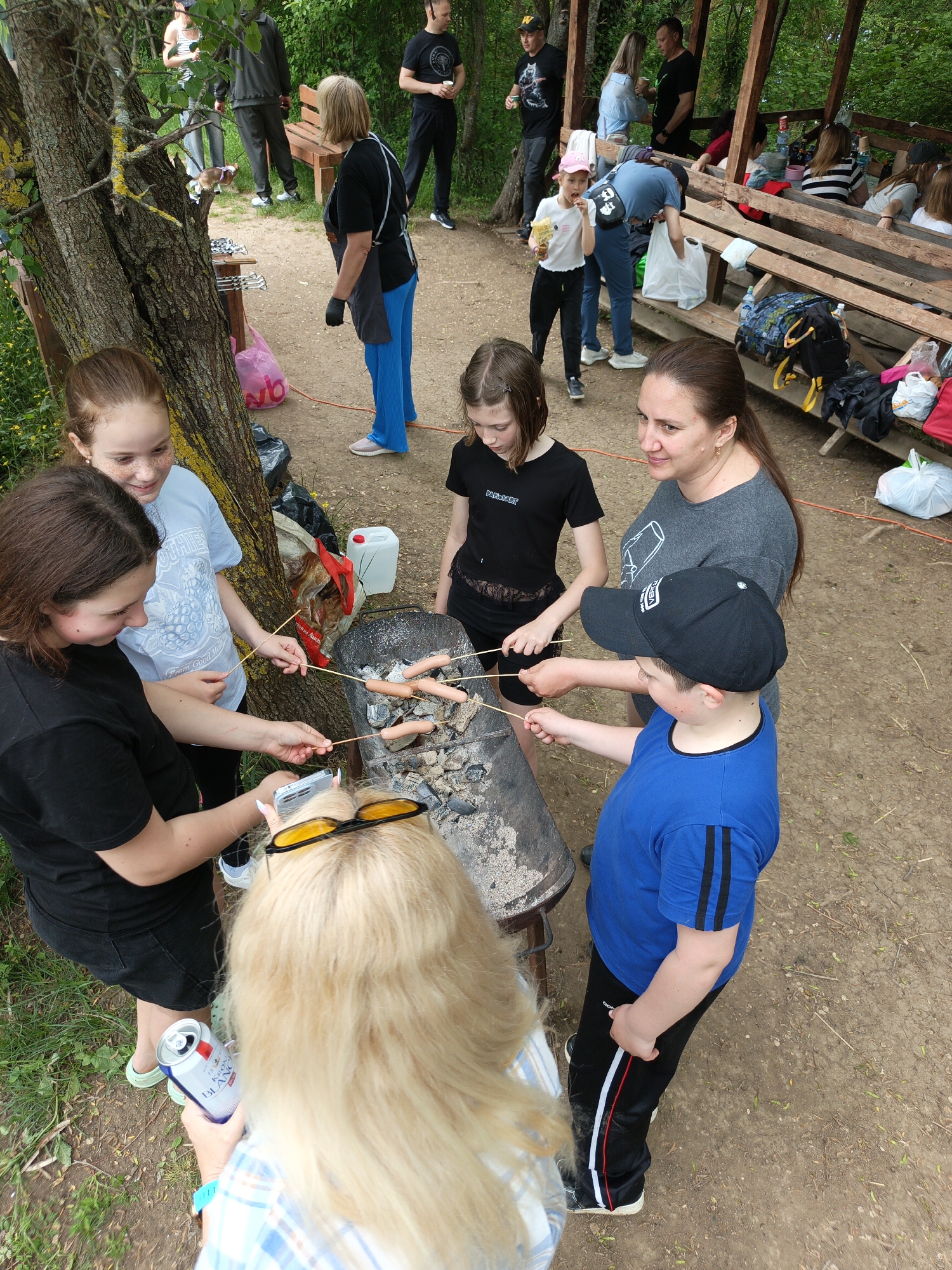 In Терновский округ, Ukraine, a group of people are gathered around a charcoal grill, roasting sausages on sticks. The group consists of a woman in a gray shirt, a boy in a blue shirt and black cap, two girls in black shirts, and another girl in a blue shirt. An individual with blonde hair and yellow sunglasses on their head is holding a can. People stand and sit in the background. A tree and a wooden structure are also visible.