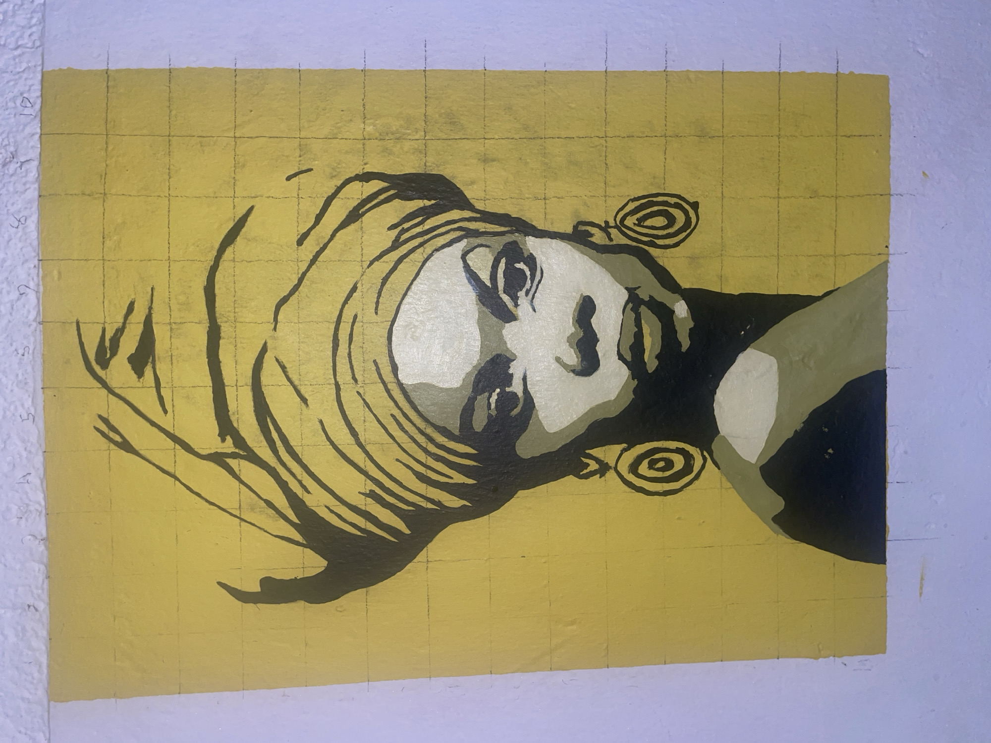 A framed painting of a woman's face, turned to the side, is affixed to a white wall. The painting features a grid pattern overlaid on a yellow background. The woman's face is rendered in shades of black, white, and grey, with abstract swirl shapes near the ears. The painting's edges are uneven and brushstrokes are visible. The scene appears to be indoors. There is no indication of a specific location within Jos, Nigeria, or of any people, actions, or interactions.