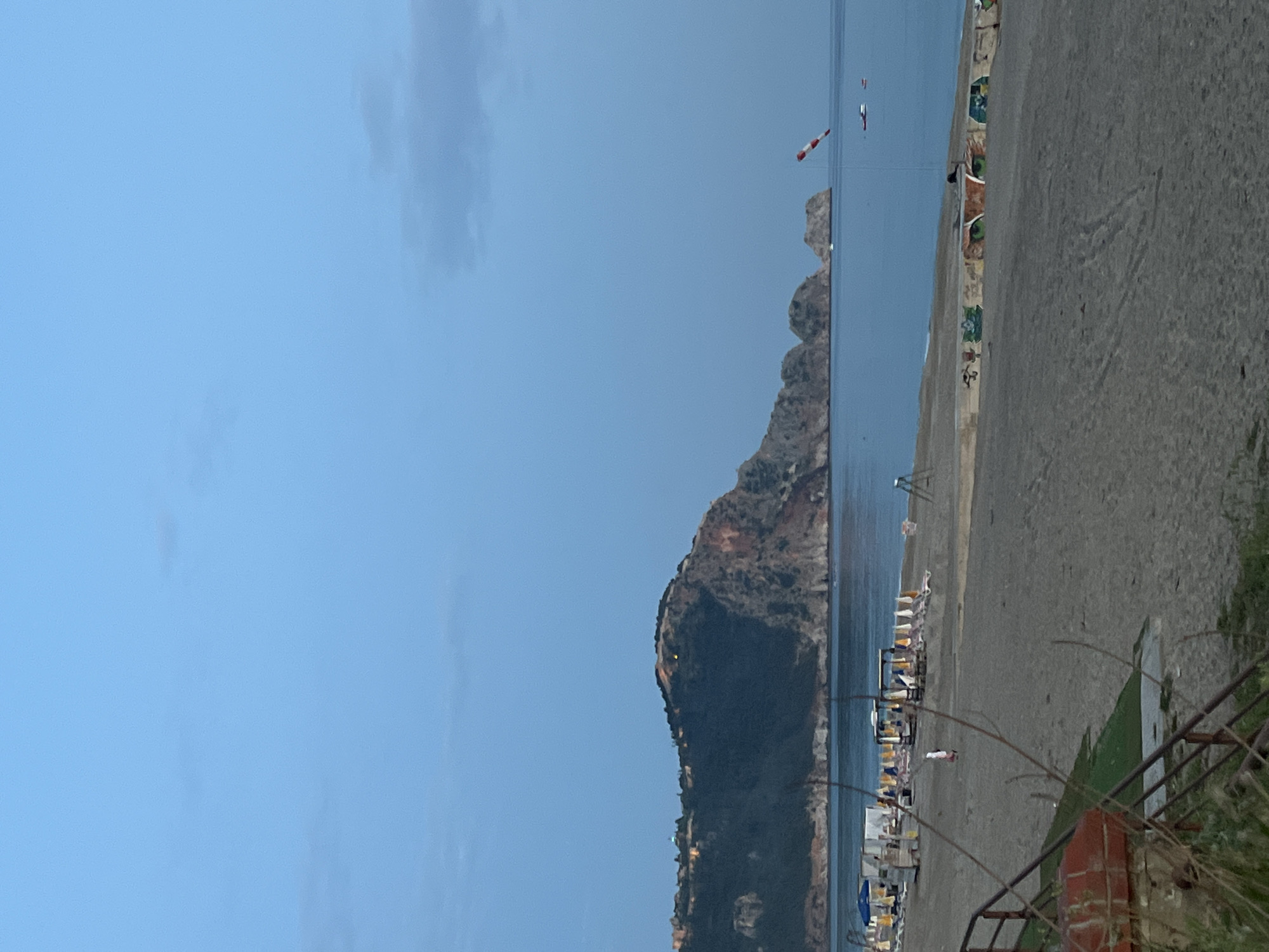 The image shows a coastal scene in Alanya, Turkey. A large, rocky mountain dominates the view, rising from the sea. A beach with numerous beach chairs and umbrellas lines the shore at the base of the mountain. The sea is a light blue, and the sky is a clear blue with a few scattered clouds. The right side of the image features a concrete wall with graffiti, indicating an urban setting near the beach. A metal railing and some foliage are in the foreground, suggesting the photo was taken from an elevated position. There are no visible people in the frame. There appears to be a small, red-and-white windsock further out in the water near another smaller rocky cliff.