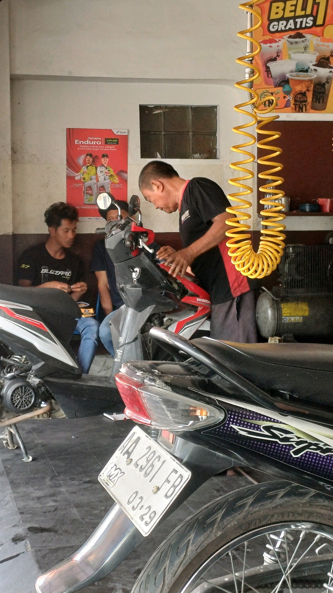The image depicts an indoor motorcycle repair shop in Temanggung, Indonesia. In the foreground, a black and silver motorcycle is visible from the rear, displaying a white license plate with the inscription "AA 2961 FB 03.29". A man in a black t-shirt with red trim is working on a red and black motorcycle, his hands manipulating its components. To his left, another man wearing a black t-shirt with the word "BLIZZARD" is seated, holding small items. Behind him, a third person is partially visible. The shop environment includes a poster on the wall featuring two men in racing attire, an advertisement for "BELI GRATIS" (Buy One Get One Free) with drinks, and a yellow coiled hose hanging from the ceiling. The walls are painted a light color, and the floor appears to be concrete. The lighting suggests an indoor setting with artificial light.