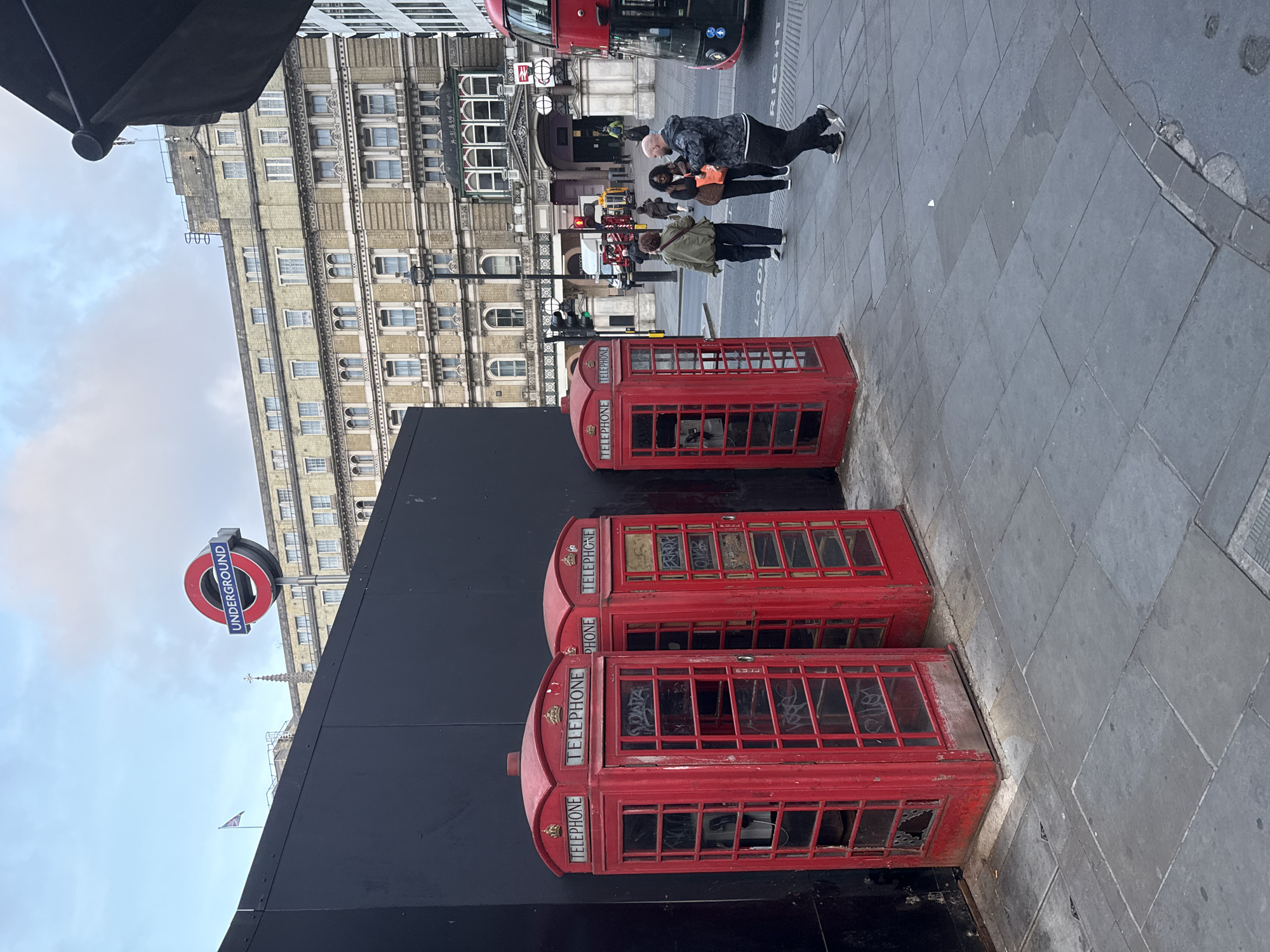The image captures a scene in London, United Kingdom, featuring three iconic red telephone boxes aligned along a sidewalk. The telephone boxes appear weathered, with graffiti visible on the glass panes. Behind the boxes is a black barrier and an Underground sign is visible in the background. Pedestrians are walking on the sidewalk, including a group of three. Buildings line the background, and a red double-decker bus is partially visible. The sidewalk is paved with gray stone.