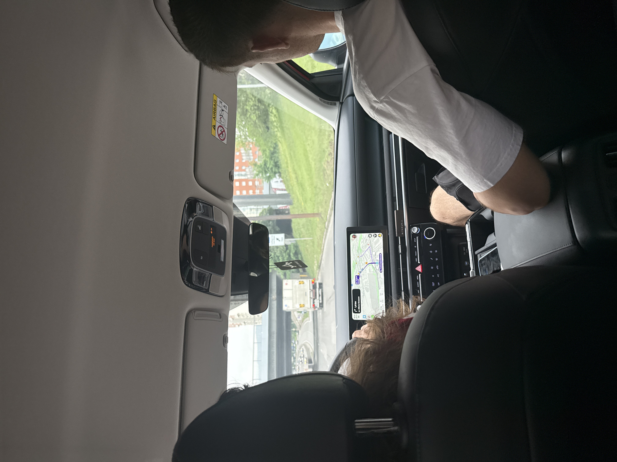 The image depicts the interior of a vehicle from the perspective of a rear passenger seat, looking forward towards the dashboard and the road ahead. In the front left, the back of the driver's head is visible, characterized by dark, curly hair. In the front right, the back of a male passenger's head, his right shoulder, and right arm are visible; he wears a white short-sleeved t-shirt and dark shorts, with his arm resting on the center console armrest.
The car's dashboard features a large infotainment screen displaying a navigation map. The map indicates a destination or nearby point of interest as "pl. Georgi Rakovski" and "ul. Vasil Levski," with a remaining distance of "1.4 km," precisely locating the scene in Sofia, Bulgaria, on a major thoroughfare heading towards the city center.
The vehicle is traveling on a multi-lane asphalt road, currently passing under a concrete overpass. Ahead, a moderate volume of traffic is observed, including a prominent white box truck in the right lane. To the right of the road, there is a grassy slope with scattered trees, behind which multi-story residential buildings with red/orange facades are visible in the distance. The sky overhead is overcast.
Inside the vehicle, details include an overhead console with light controls and a "PASSENGER AIR BAG ON" indicator. The passenger-side sun visor is down, revealing an "AIRBAG" warning label with pictorial instructions. A black air freshener with a white stylized 'A' logo hangs from the rearview mirror. The center console also shows branding for "RITUALS..." below the climate controls.