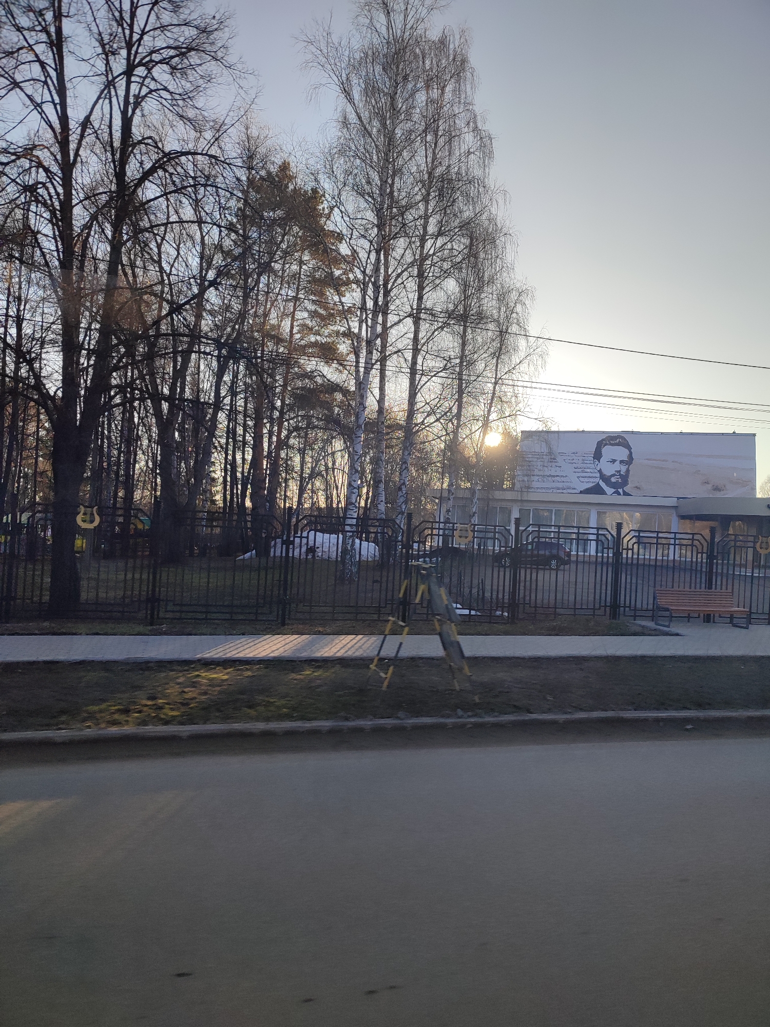 The image shows an exterior view in what is probably Chaikovsky, Russia, based on the mural of Pyotr Ilyich Tchaikovsky on the building. A black wrought-iron fence runs across the midground, separating a sidewalk and street from the grounds of a building. Bare trees are interspersed behind the fence. Behind the fence, there is a mural of Tchaikovsky, a building, vehicles, and trees. A yellow ladder is propped on the near side of the fence. The street comprises the foreground. The sky is overcast, with sunlight peeking through the trees.