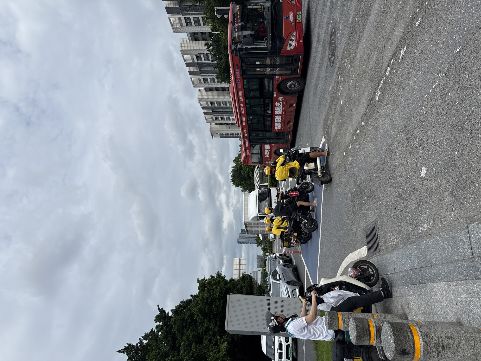 In Haizhu District, China, a group of individuals on electric scooters, some wearing yellow uniforms, are stopped at an intersection. A large red bus is in motion on the right side of the frame, and a white truck is visible behind it. Several cars are also present, including a white sedan on the left. The scene is urban, with modern buildings in the background and trees lining the left side of the image. The sky is overcast with gray clouds.