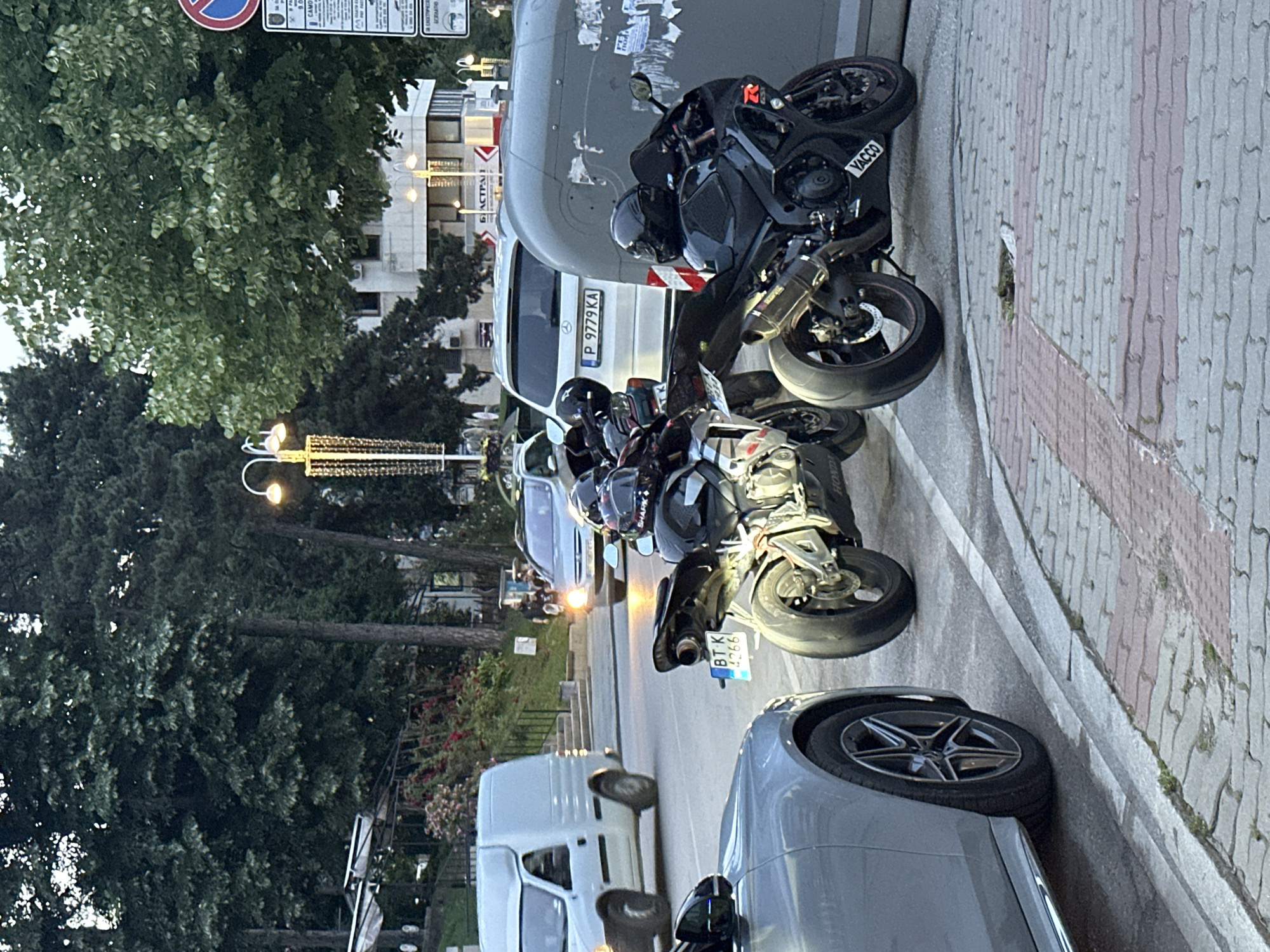 The image, taken in Veliko Tarnovo, Bulgaria, captures a street scene during what appears to be late afternoon or early evening, characterized by soft ambient light and illuminated street lamps. The primary subjects are several motorcycles and cars parked alongside a paved sidewalk.
In the foreground and center, at least three motorcycles are prominently displayed, parked on the sidewalk. One black sportbike features "YACCO" branding on its side fairing and a helmet with "SHARKX" text resting on it. Another motorcycle, partially visible, has a Bulgarian license plate "BT K 4266", with "BT" confirming its registration in the Veliko Tarnovo region.
Behind the motorcycles, a white Mercedes-Benz van is parked, displaying the Bulgarian license plate "P 9779 KA", indicating registration in the Plovdiv region. To the far right, part of a dark grey car is visible. The street itself appears busy, with other vehicles and bright headlights seen in the distance.
The background is rich with green foliage from numerous trees, suggesting a verdant urban environment. Beyond the trees, buildings with illuminated windows and a banner bearing Cyrillic text, "Б АСТРАЛ", are discernible. Street lights with ornate fixtures cast a warm glow. A circular red and blue road sign, likely indicating a prohibition, is visible in the upper left. The sidewalk is paved with grey and reddish-brown interlocking bricks. No direct human activity is evident, but the parked motorcycles with helmets suggest their riders are nearby.