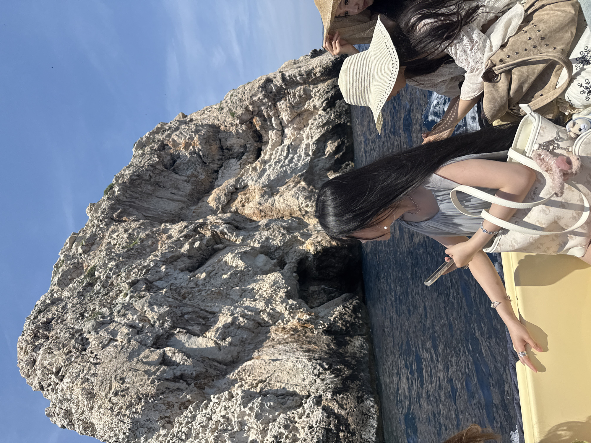 In the city of Komiža, Croatia, on a boat trip near a rocky cliff face with a cave opening, three women are captured in a medium shot. The woman on the left, facing away from the camera, has long, dark hair and wears a light blue dress, a silver necklace, and a heart-shaped charm bracelet. Her left hand rests on the yellow boat railing, adorned with a silver ring, while her right hand holds a clear phone case. Beside her, a woman in the center wears a wide-brimmed straw hat and a white lace top, with a tan suede purse at her side. Partially visible on the far right, another woman is seen wearing a straw hat. The boat, with its yellow railing, is on a dark blue body of water under a bright blue sky.