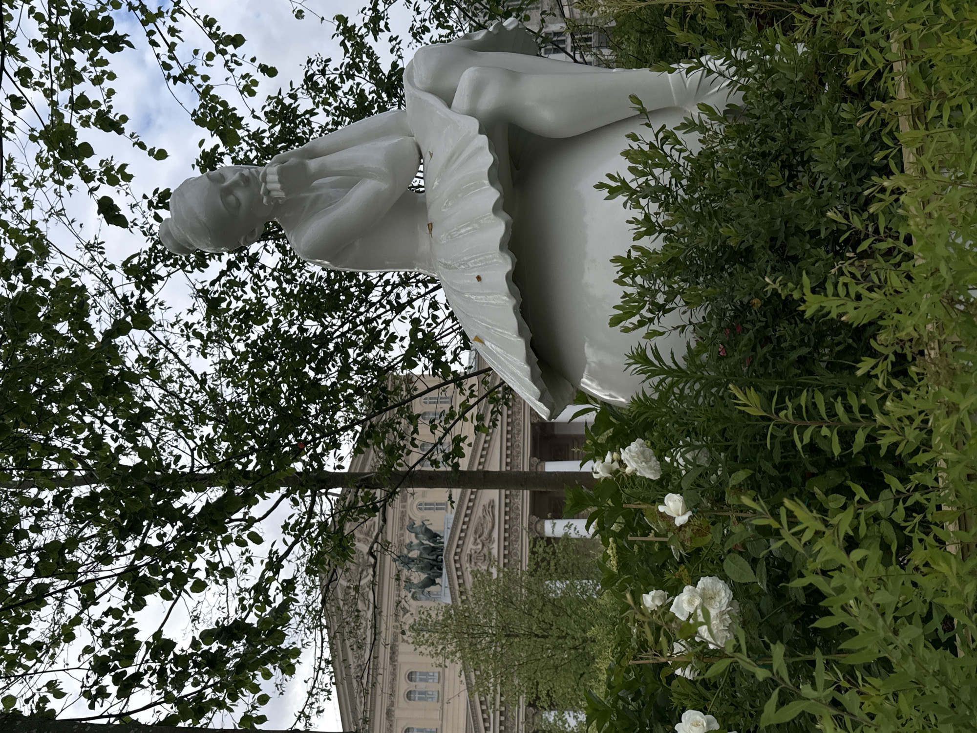 The image depicts a white statue of a ballerina in a tutu, placed within a garden setting. White roses bloom around the statue. In the background, the Bolshoi Theatre in Moscow, Russia, is visible. The composition includes trees and foliage, and the sky is partly cloudy.