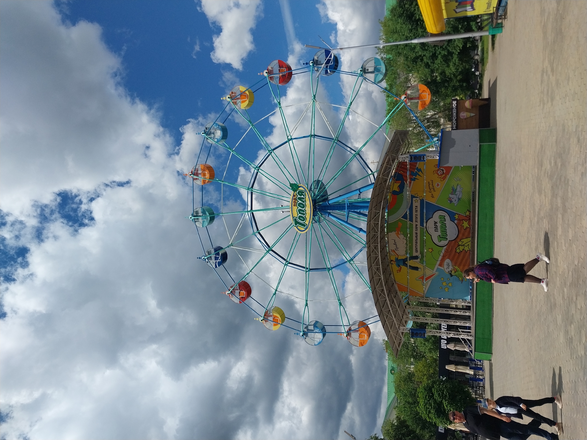 The image captures a vibrant scene at a park in Orenburg, Russia. The dominant feature is a colorful Ferris wheel with gondolas in shades of orange, yellow, blue, and red, set against a backdrop of a partly cloudy blue sky. To the right of the Ferris wheel, there is a structure with a colorful cartoon-like mural, adorned with the name of the park "Тополя" (Topolya) in Russian.
A few people are present: one person is standing and facing the mural, and two people are walking away. The environment includes trees, paving stones, and other park elements. The scene appears to be taken on a sunny day.
