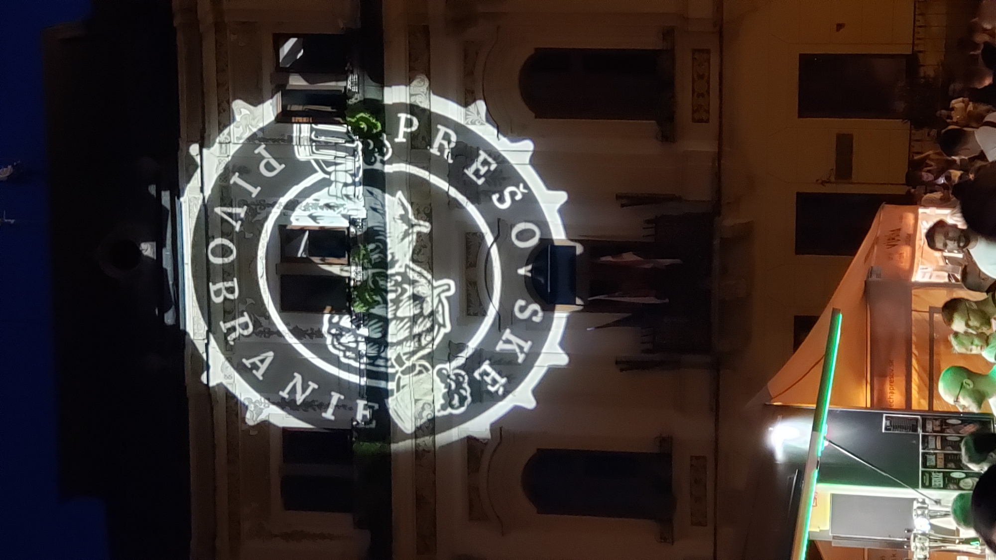 An exterior night shot captures a building in Prešov, Slovakia, illuminated by a projector casting a circular logo onto its facade. The logo appears to be a stylized beer bottle cap with text around the perimeter. The building has arched windows and decorative architectural elements. In the foreground, a crowd of people are gathered in front of food and drink stalls, suggesting an outdoor event or festival. One stall has green LED lighting. The overall atmosphere is lively and celebratory.