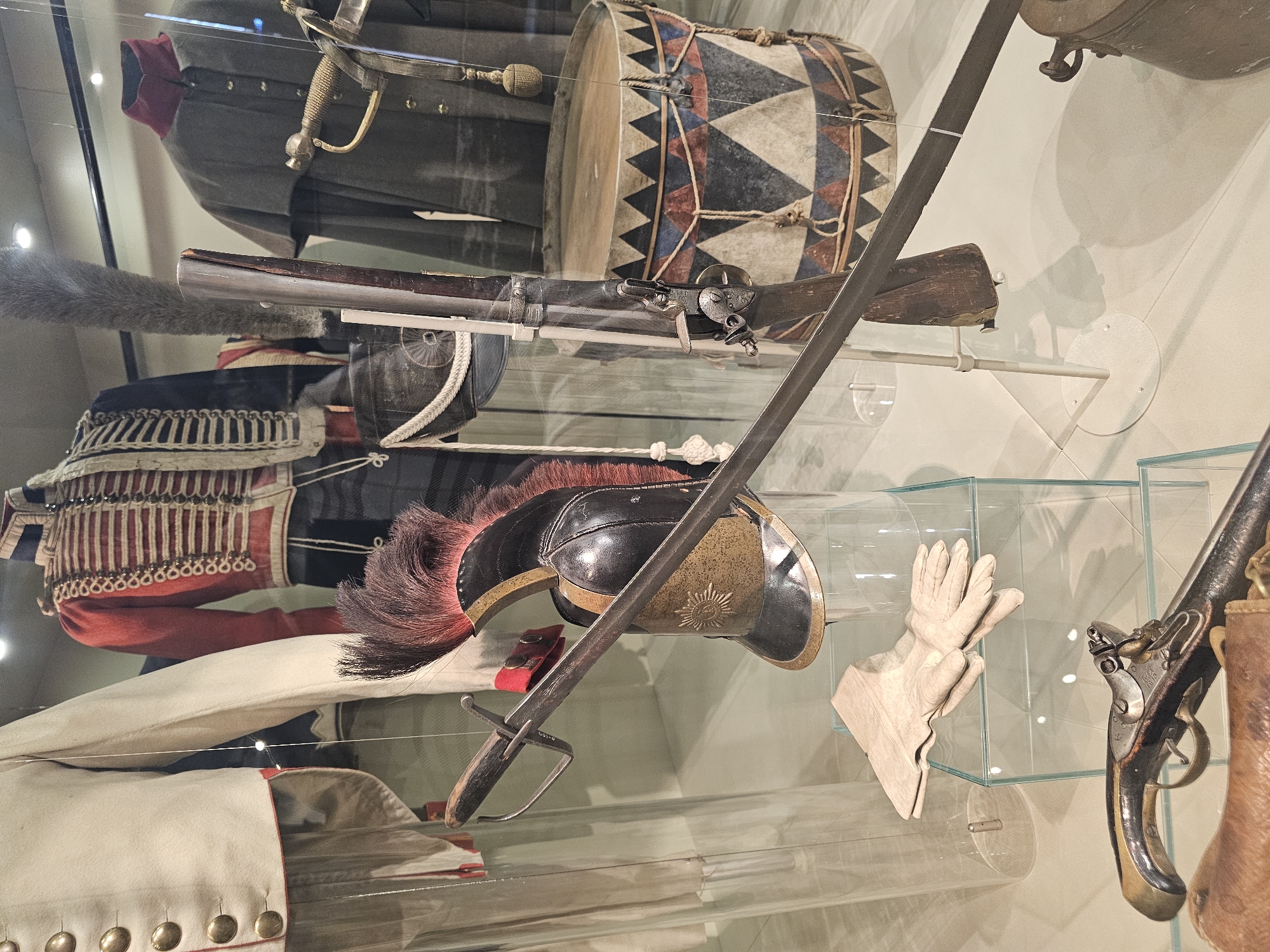 The image shows a display case in a museum, likely related to military history, potentially located in Borodino, Russia, given the subject matter. The display features a collection of artifacts, including military uniforms in various colors (white, red, black, and grey), indicating different regiments or ranks. A large drum with a geometric pattern is present. Firearms, including a musket and a pistol, are also displayed. A sword and a helmet with a plume add to the military theme. A pair of leather gloves are also included in the display. The environment is sterile, with the items presented behind glass on transparent supports.