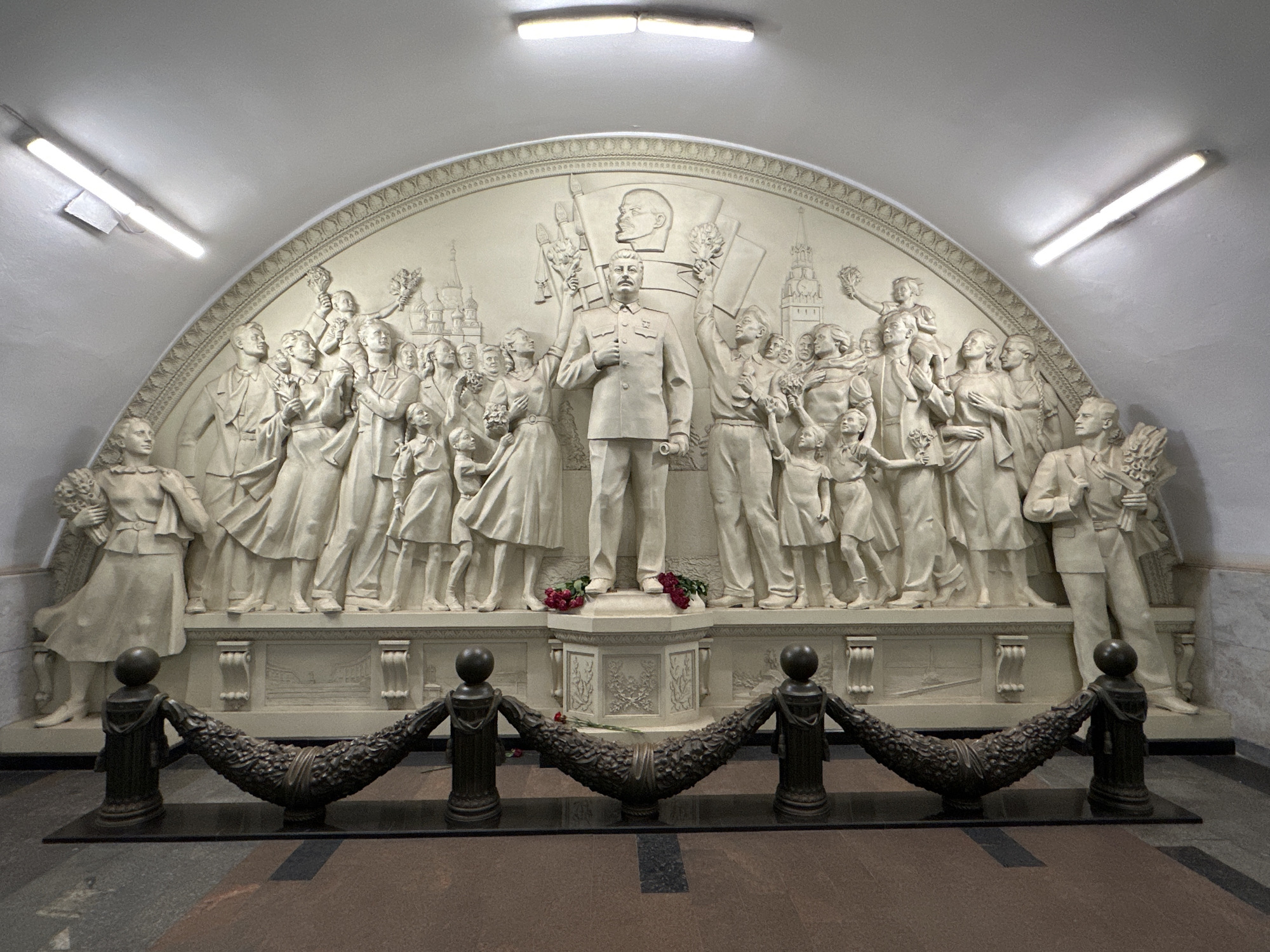 The image shows a sculptural composition in the Moscow Metro, Russia. At the center is a high-relief sculpture of a man in a military-style suit, likely representing Joseph Stalin, surrounded by a crowd of figures, including adults and children, with a depiction of Lenin in the background. The figures are depicted holding flowers and banners, suggesting a celebratory or propagandistic scene. The sculpture is set within an arched niche adorned with decorative molding. Below the sculpture, there is a barrier consisting of posts with decorative garlands. The floor is tiled. The lighting in the scene is provided by fluorescent lights on the ceiling, illuminating the sculpture.