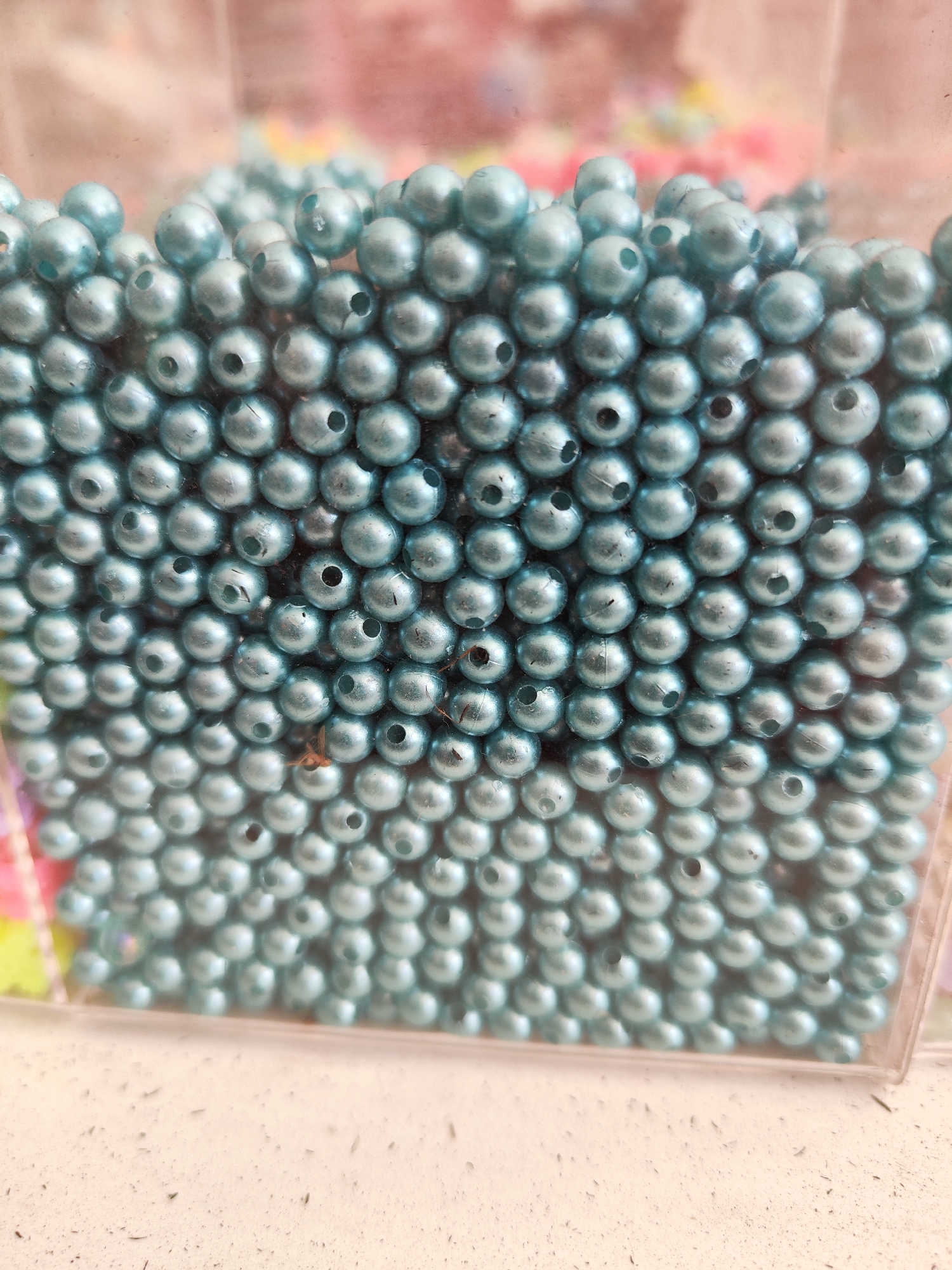 The image shows a close-up, eye-level view of a transparent container filled with numerous small, iridescent teal beads. The beads are spherical and appear to have a hole through their center, suggesting they are intended for crafting or jewelry making. They are densely packed within the container, creating a textured surface with varying depths. The lighting highlights the pearlescent sheen of the beads, causing some to appear brighter than others. In the blurred background, other colorful beads or craft materials are visible, indicating a crafting supply context. The container is resting on a light-colored, speckled surface, possibly a table or shelf. The image does not depict any individuals or specific locations within Sragen, Indonesia.
