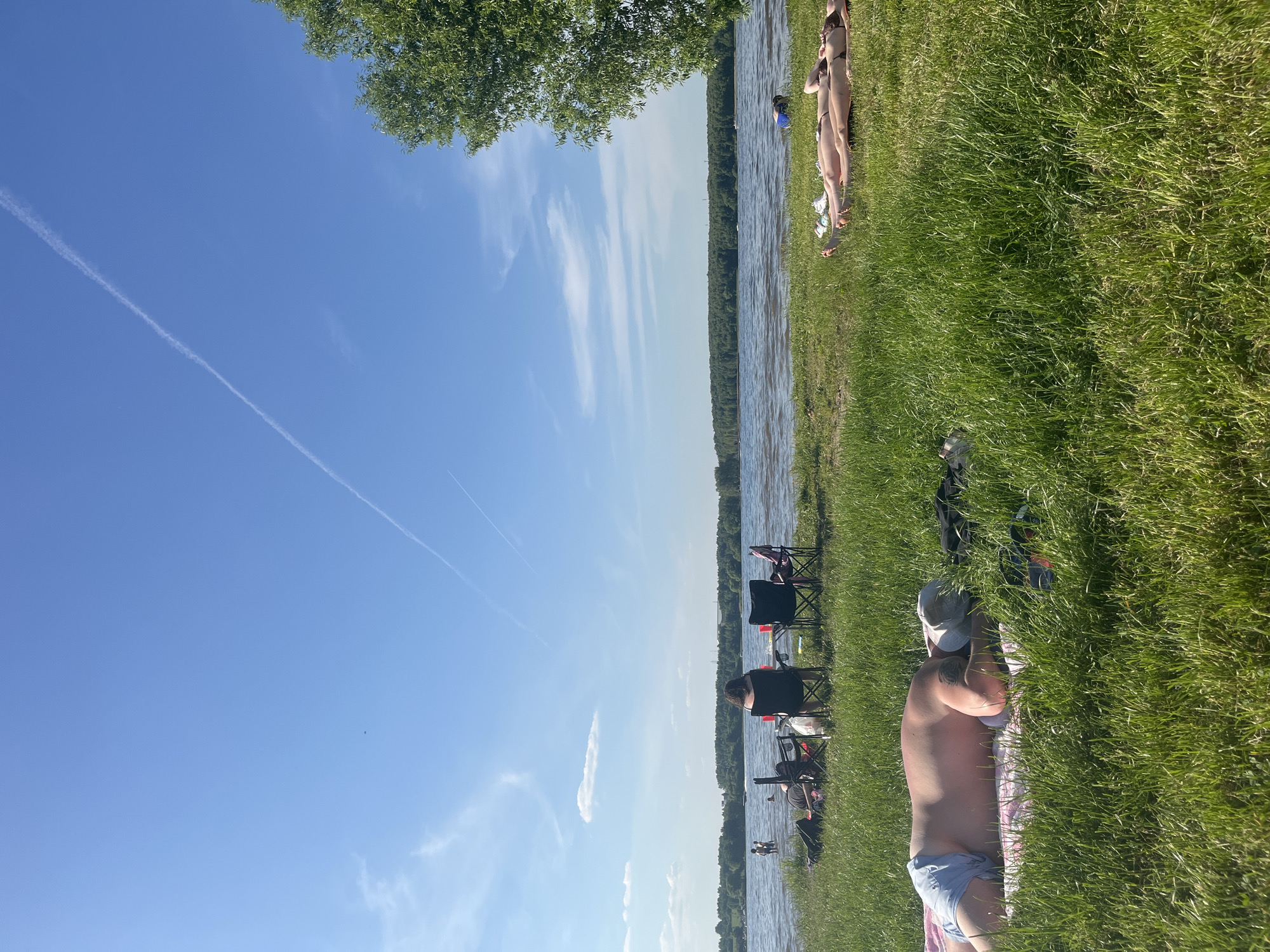 This detailed description pertains to an image captured on a sunny day at a recreational area in Kapustino, Russia. The scene depicts a wide body of water, likely a lake or reservoir, with a grassy shoreline in the foreground and a dense forest lining the distant bank. The sky is a clear blue, adorned with multiple white aircraft contrails and a few scattered, wispy cumulus clouds.
In the immediate foreground, towards the left, a person, appearing male, lies face down on a pink towel placed on vibrant green grass. He wears light blue shorts and a light-colored baseball cap on his head, with his back and shoulders exposed, showing some sun-reddening and a dark tattoo on his upper right arm.
Mid-ground, closer to the water's edge, a group is situated. One individual, with dark hair, is seated in a dark folding chair, facing the water with their back to the camera. Adjacent to this person is a small folding table holding two red cups and an empty dark folding chair. Bags and personal belongings are scattered on the grass around these chairs. In the water directly in front of this group, several individuals are visible, either wading or swimming.
Further to the right in the mid-ground, two individuals are lying prone on a light-colored blanket or towel on the grass, sunbathing. They are facing away from the camera, wearing swimsuits, and positioned near the base of a large green tree that frames the right side of the image.
The water surface displays ripples, indicating a light breeze. In the distance, faint outlines of structures or boats can be discerned on or near the far forested bank. The overall environment suggests a tranquil, leisure-oriented outdoor setting under clear, bright daylight.