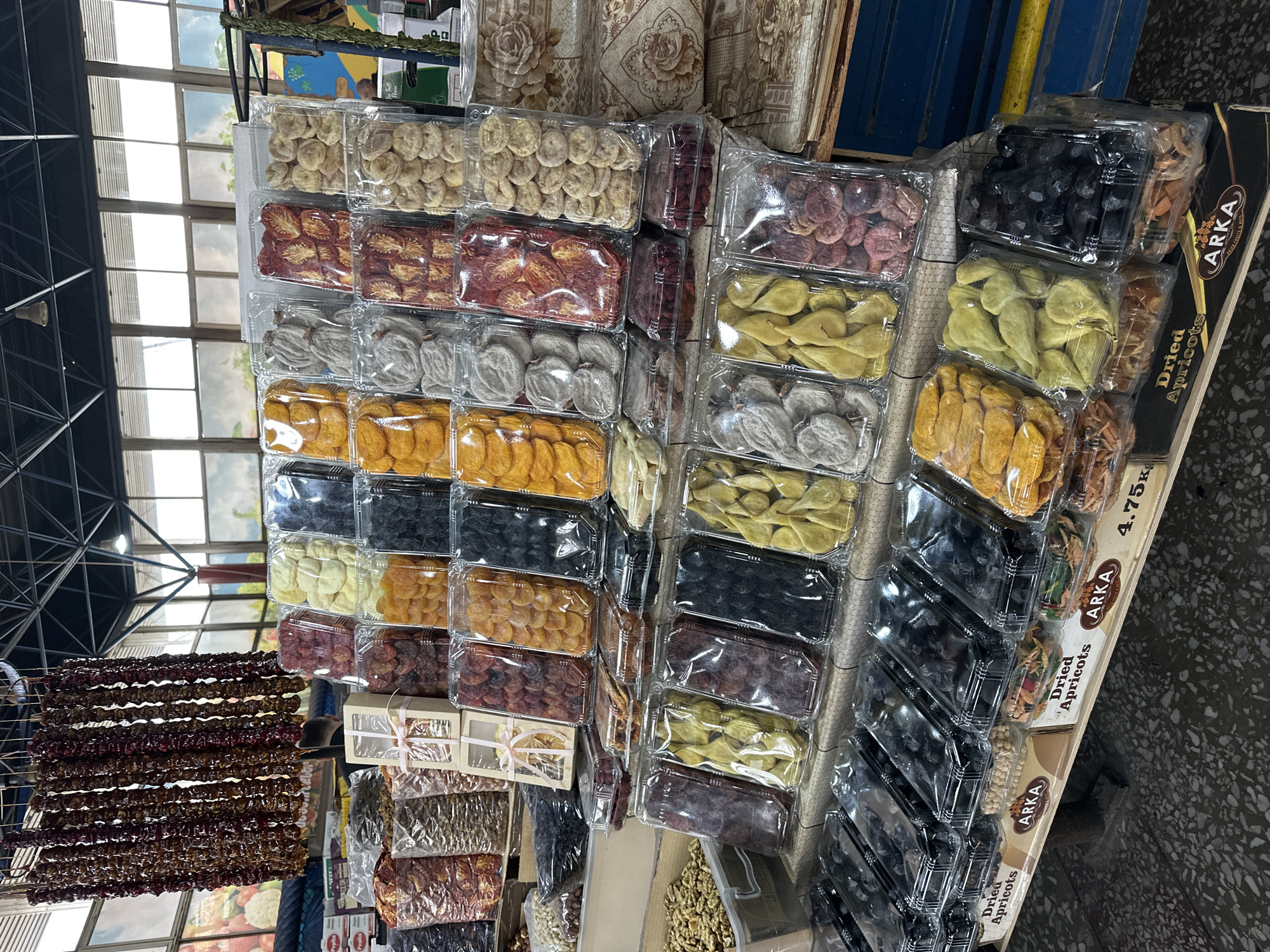 The image captures a display of dried fruits and sweets in Yerevan, Armenia. Trays and boxes of various dried fruits such as apricots, figs, persimmons, pears, and plums are arranged for sale. A long hanging string of a traditional sweet snack known as "churchkhela," made of nuts dipped in thickened fruit juice, is visible in the lower left corner. Branded packaging indicates some products are "Dried Apricots" and labeled "ARKA". Other nuts such as walnuts, pistachios and seeds, are also on display. The setting appears to be an indoor market or shop.