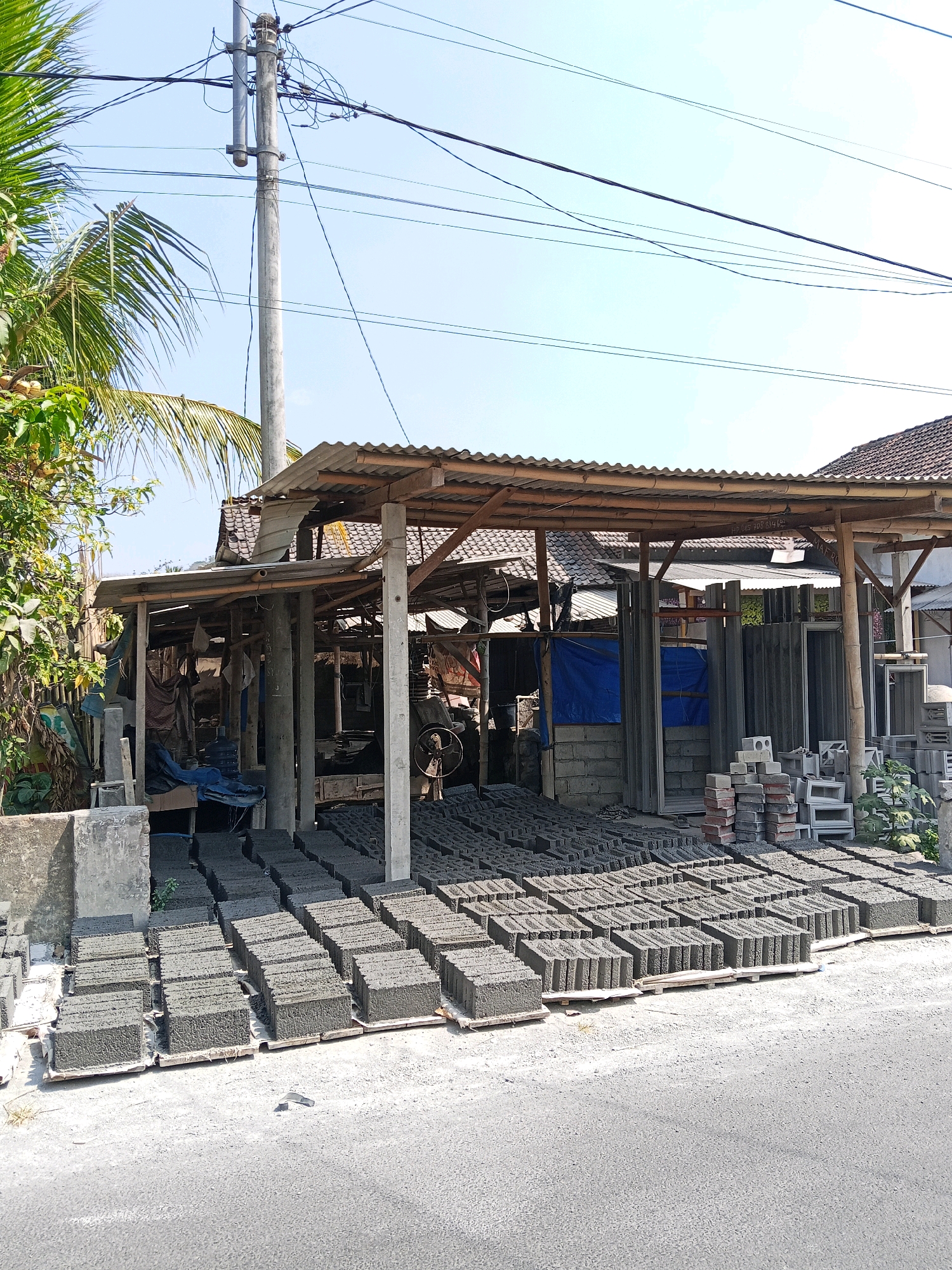 This is an outdoor, eye-level, medium shot of a brick-making facility in Margo Mulyo, Indonesia. The scene is dominated by rows of concrete bricks laid out on wooden pallets, neatly arranged on a gravel surface. Behind the bricks, a simple, open-sided structure with a corrugated metal roof, supported by wooden beams, houses a brick-making machine, various supplies, and some blue tarps. The background shows a tiled roof of a building and a utility pole with numerous wires. A palm tree and other greenery are visible to the left of the frame, and the ground in the foreground is paved with asphalt. The sky is clear and blue.