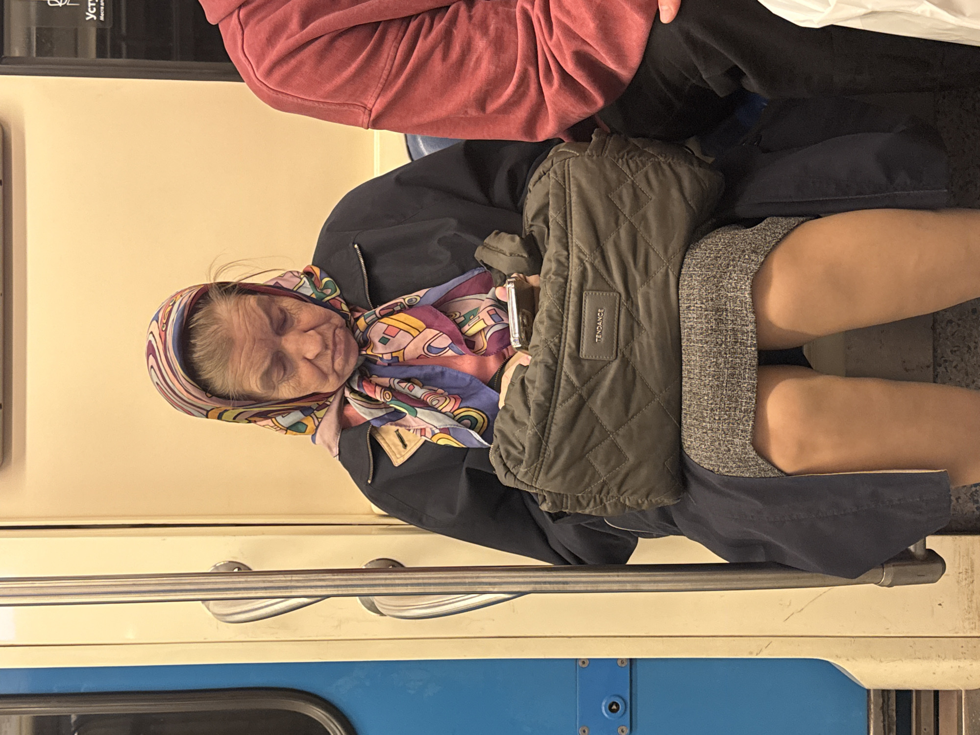 An elderly woman with a colorful headscarf is asleep on a train, presumably in Moscow, Russia, based on the interior. She is wearing a dark blue jacket and a grey skirt, with a quilted olive-green bag on her lap. A man in a red sweater sits beside her. The train car is blue and cream-colored with metal handrails.