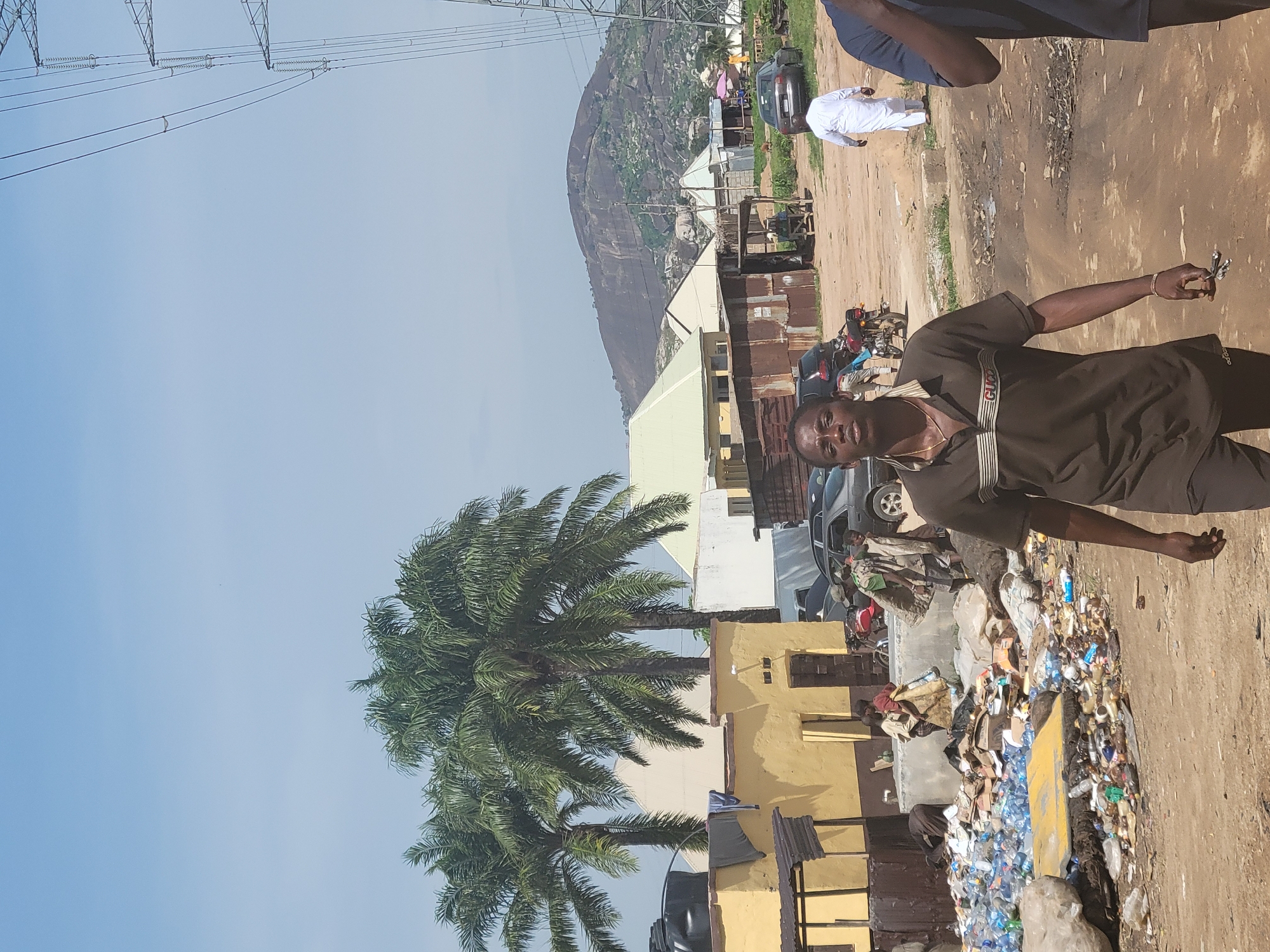 The image depicts a scene in Kwamba, Nigeria, characterized by a dirt road and a significant amount of accumulated garbage, primarily plastic bottles and cardboard. In the foreground, a man wearing a brown polo shirt and dark shorts walks towards the camera. He has a gold chain around his neck and is holding a small bunch of keys in his right hand. In the middle ground, several people are visible. Some are carrying sacks, and others are near vehicles. A person in a white garment walks away from the camera in the background, and another person in a blue shirt is partially visible on the right edge of the frame. The environment includes palm trees, residential structures with yellow and white walls, corrugated metal sheds, and parked cars and motorcycles. In the distance, a large, rocky hill dominates the horizon under a clear sky. Power lines and a transmission tower are also present in the frame.