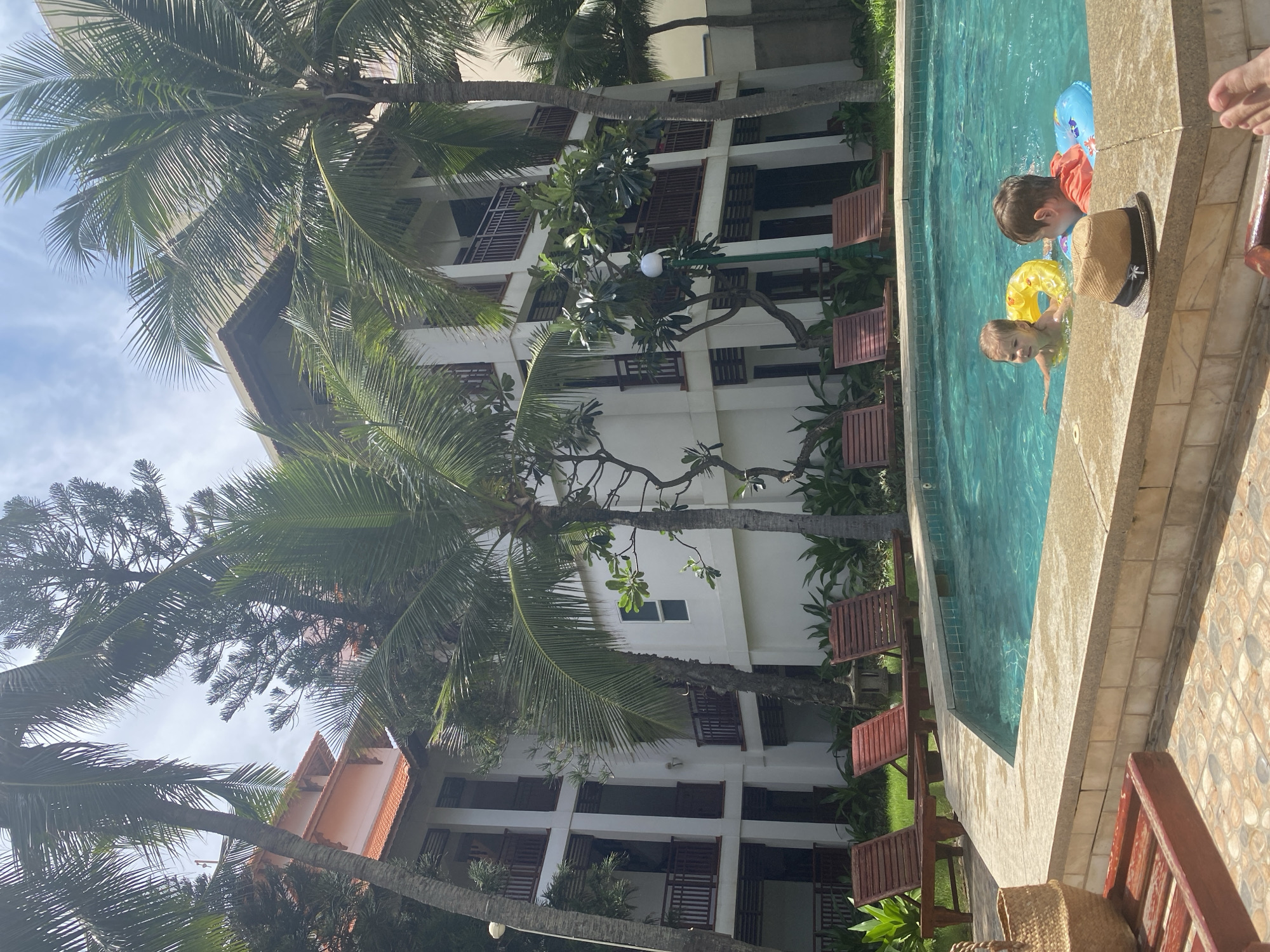 Two young children, a girl and a boy, are playing in a swimming pool at a resort in Phu Thuy Ward, Vietnam. The girl is wearing a yellow inflatable ring and the boy is wearing a blue inflatable ring. They are both looking towards the camera. In the foreground, a straw hat rests on the edge of the pool. In the background, a multi-story white building with balconies is visible, surrounded by palm trees. Several lounge chairs are arranged around the pool. The sky is cloudy.