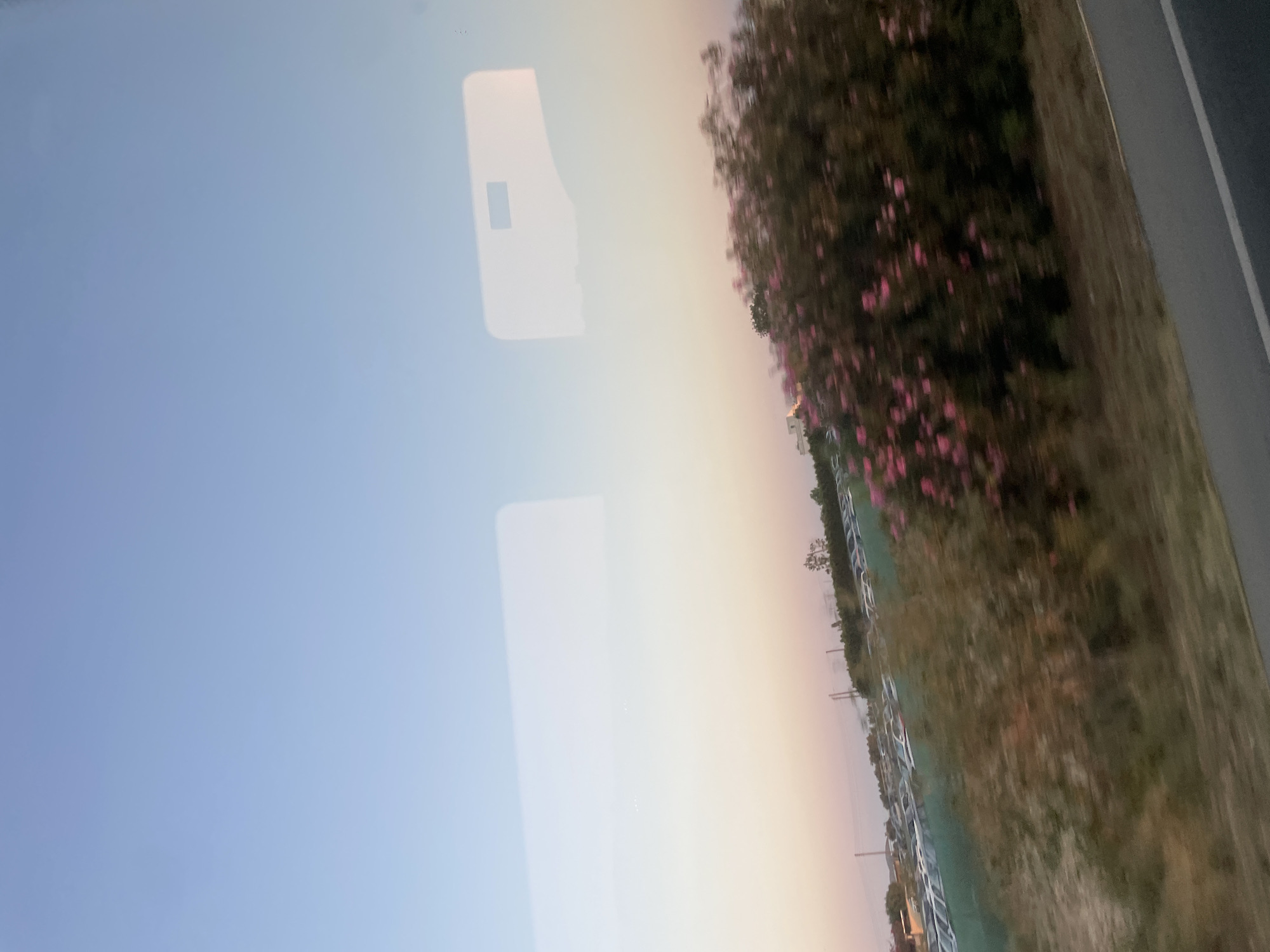 The image depicts a scene from a vehicle, likely a bus or car, looking out the window. The dominant element is the sky, which transitions from a pale blue at the left to a soft, warm gradient of yellow and pink towards the right, suggesting either sunrise or sunset.
To the right of the sky, a blurry line of vegetation is visible, featuring dark green foliage interspersed with splashes of pink flowers. This vegetation appears to be along the side of a road or an open area. Further to the right, a grey, asphalt-like surface suggests a road or pathway, with a thin white line marking its edge. In the lower right corner, the scene is darker, hinting at the approach of evening or early morning.
Reflected on the window are bright, rectangular shapes, possibly from the interior of the vehicle. One prominent reflection, slightly left of center, is a large, pale rectangle with a smaller, darker rectangle inside it, resembling a window or a display screen. There are also faint, shimmering lights scattered across the sky portion of the reflection.
The overall impression is one of travel through a landscape during a transitional time of day, with the natural beauty of the sky and flowering plants contrasting with the man-made elements of the road and the vehicle's interior. The time of day is either dawn or dusk, and the weather appears clear and calm, based on the soft light and lack of atmospheric disturbance. While the location is identified as Timi Community, Cyprus, there are no specific landmarks or text to further pinpoint the exact location within the community.