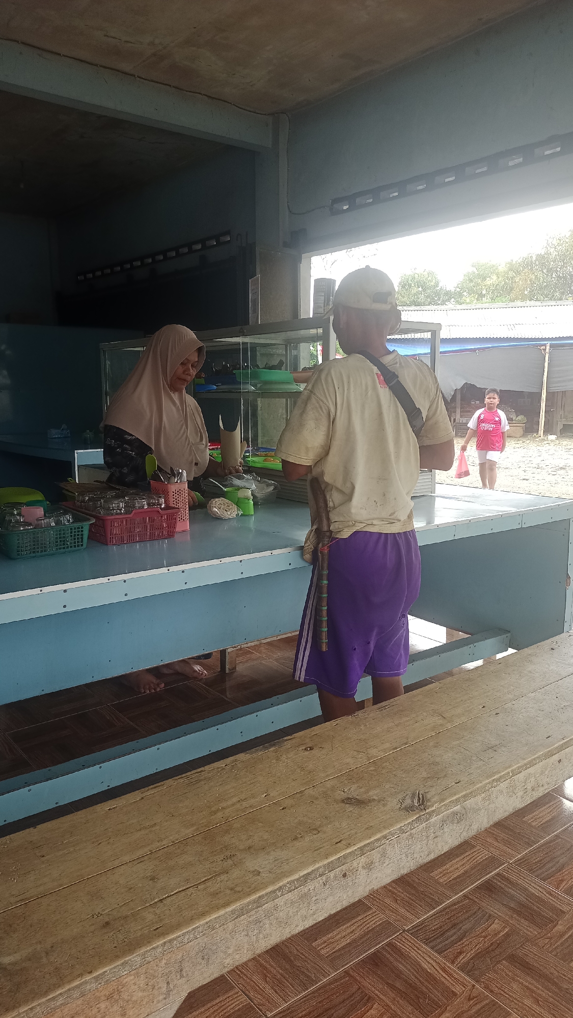 A woman wearing a light brown hijab and a patterned top stands behind a blue counter in what appears to be an outdoor food stall or small restaurant. She is facing left, attending to a display case. To her right, a man wearing a light yellow t-shirt, dark shorts, and a baseball cap stands facing away from the camera, looking at the woman. He has a dark strap across his chest and what appears to be a wooden object tucked into his waistband. In the background, a younger boy in a pink and white shirt walks away from the stall, carrying a pink plastic bag. The environment is simple, with a concrete ceiling, blue counter, and tiled floor. The scene is set in Karyamandala, Indonesia.