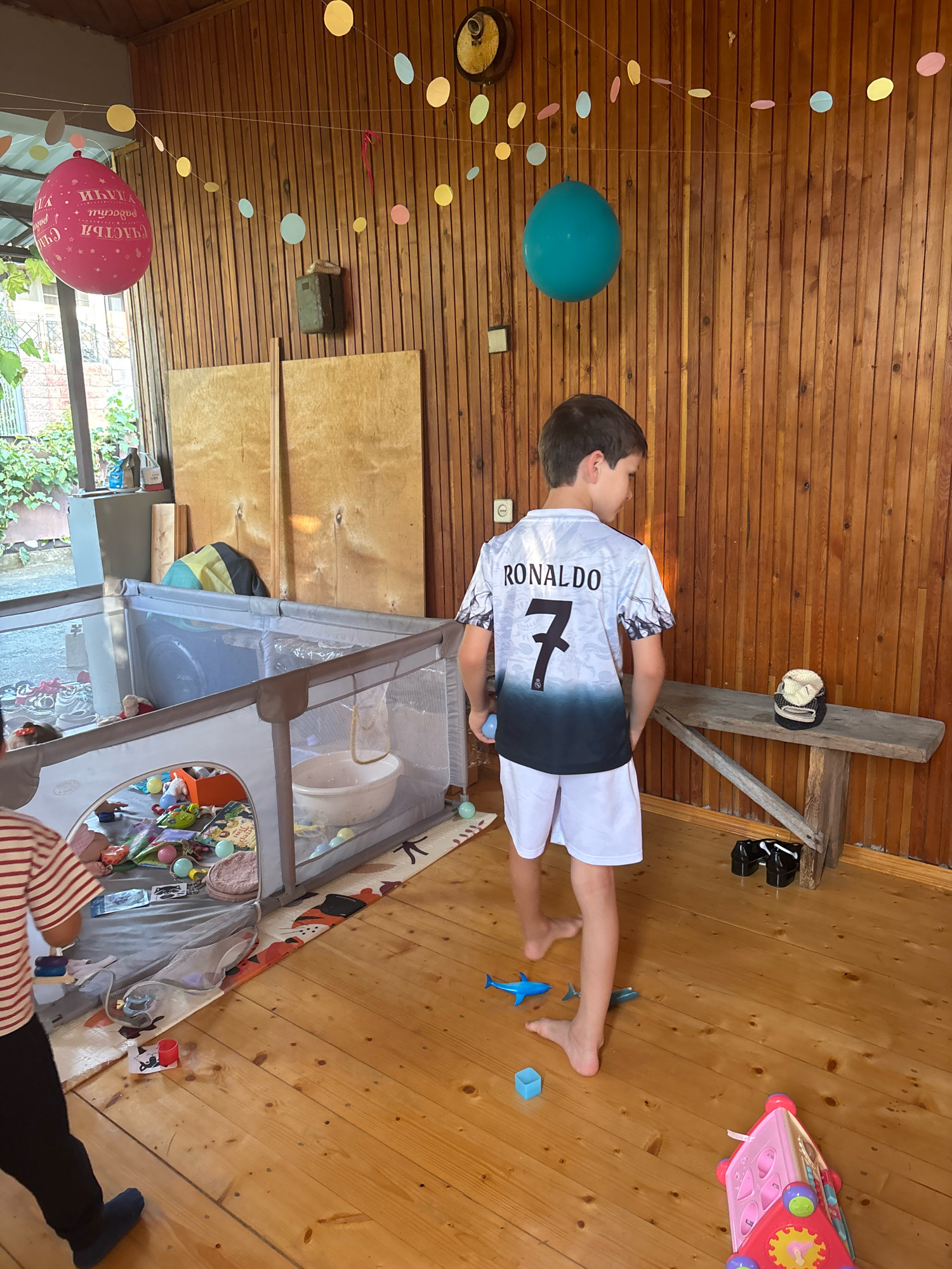 The image depicts an indoor scene, possibly a covered patio or porch, with wooden walls and a wooden floor. The space is decorated with balloons and paper garlands, suggesting a celebration. In the foreground, a young boy wearing a soccer jersey with "RONALDO 7" on the back and white shorts stands facing away from the camera. To the left, another child, partially visible, is wearing a striped shirt and black pants, and appears to be standing near a playpen filled with toys. The playpen contains various items including soft balls, blocks, and stuffed animals. A wooden bench with a cap on it is visible against the back wall. Other objects in the scene include a teal balloon, a pink balloon, and several small toy sharks on the floor. The lighting suggests it is daytime. The location is identified as Unknown, Unknown.