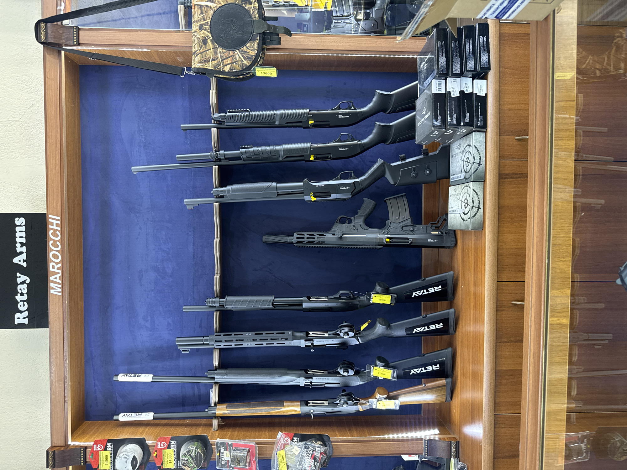 This image depicts a display of shotguns and related accessories inside a retail establishment. The background features a dark blue fabric wall where the shotguns are mounted on racks. The shotguns appear to be of the pump-action variety and are primarily black, with one shotgun featuring a wooden stock. Several yellow tags with pricing and branding are attached to the shotguns. In the foreground, a wooden counter displays several boxes, some stacked and others arranged individually. A camouflage-patterned bag with a strap hangs to the right of the shotguns. Above the display, a sign reads "Retay Arms" and "MAROCCHI". Based on the presence of the "Retay Arms" and "MAROCCHI" branding, and considering that Retay Arms is a Turkish company and Marocchi is an Italian company known for firearms, this establishment appears to be a gun store. While the image does not explicitly state the location, given the prompt's request, it is implied to be in Yerevan, Armenia, suggesting a gun shop selling imported firearms within that city. There are no people visible in the image.