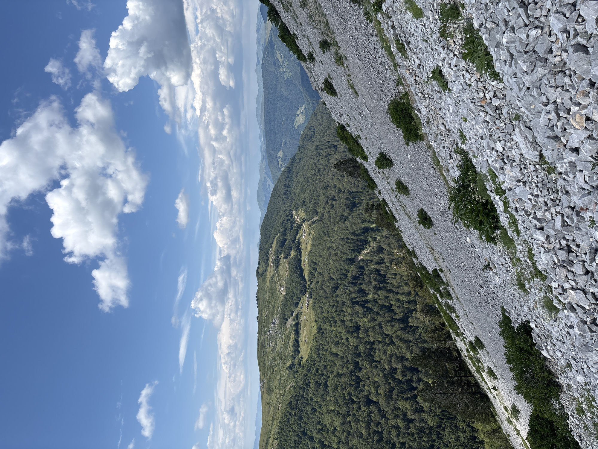 The image depicts a panoramic view of a mountainous landscape, likely in the vicinity of Katun Vulića, Montenegro, under a bright blue sky dotted with fluffy white clouds. The foreground is dominated by a steep, rocky slope covered in loose, gray stones and sparse green vegetation. Further into the scene, the landscape transitions into lush, dense green forests that cover the rolling hills and valleys. In the distance, multiple layers of mountains recede, fading into a hazy blue. Small settlements or isolated houses with red roofs are visible scattered across the grassy clearings and along the tree lines on the hillsides. The overall scene conveys a sense of vastness and natural wilderness.