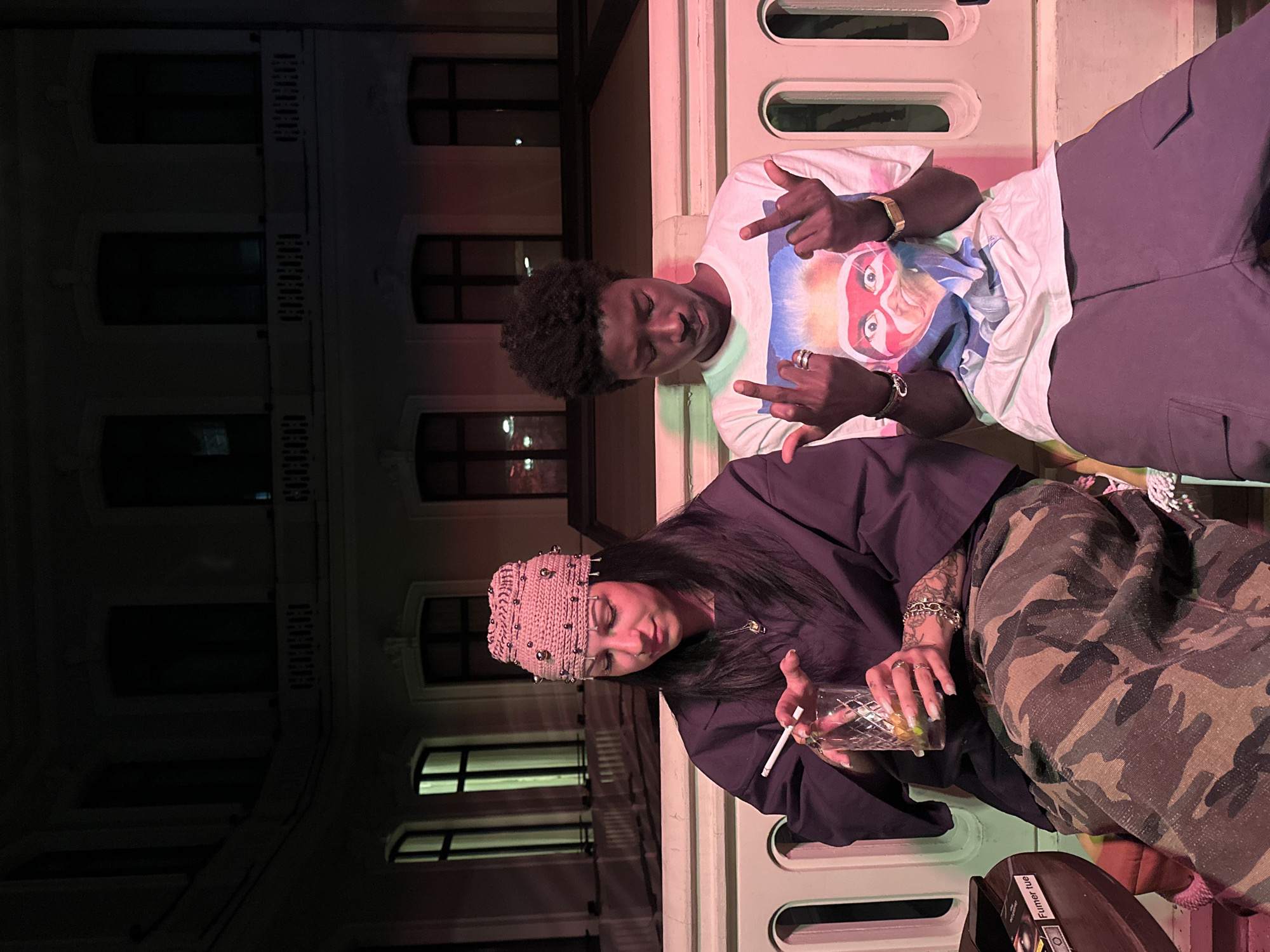 The photograph depicts two individuals, a man and a woman, seated outdoors in what appears to be a relaxed setting. The man, positioned towards the upper part of the frame, is wearing a white t-shirt with a graphic print, and grey trousers. He has dark skin and curly hair, and is making a hand gesture. The woman, seated lower in the frame, is wearing a dark top, camouflage trousers, and a studded knitted beanie. She is holding a glass with a beverage and a cigarette.
The background shows a light-colored building with windows. The location appears to be in Paris, France. The presence of a "Fumer tue" (Smoking kills) sticker on a nearby object further indicates a European setting. The individuals are seated next to a structure with rounded, elongated openings, suggesting a balcony or elevated outdoor area. The scene is illuminated with pink and green tinged artificial light.