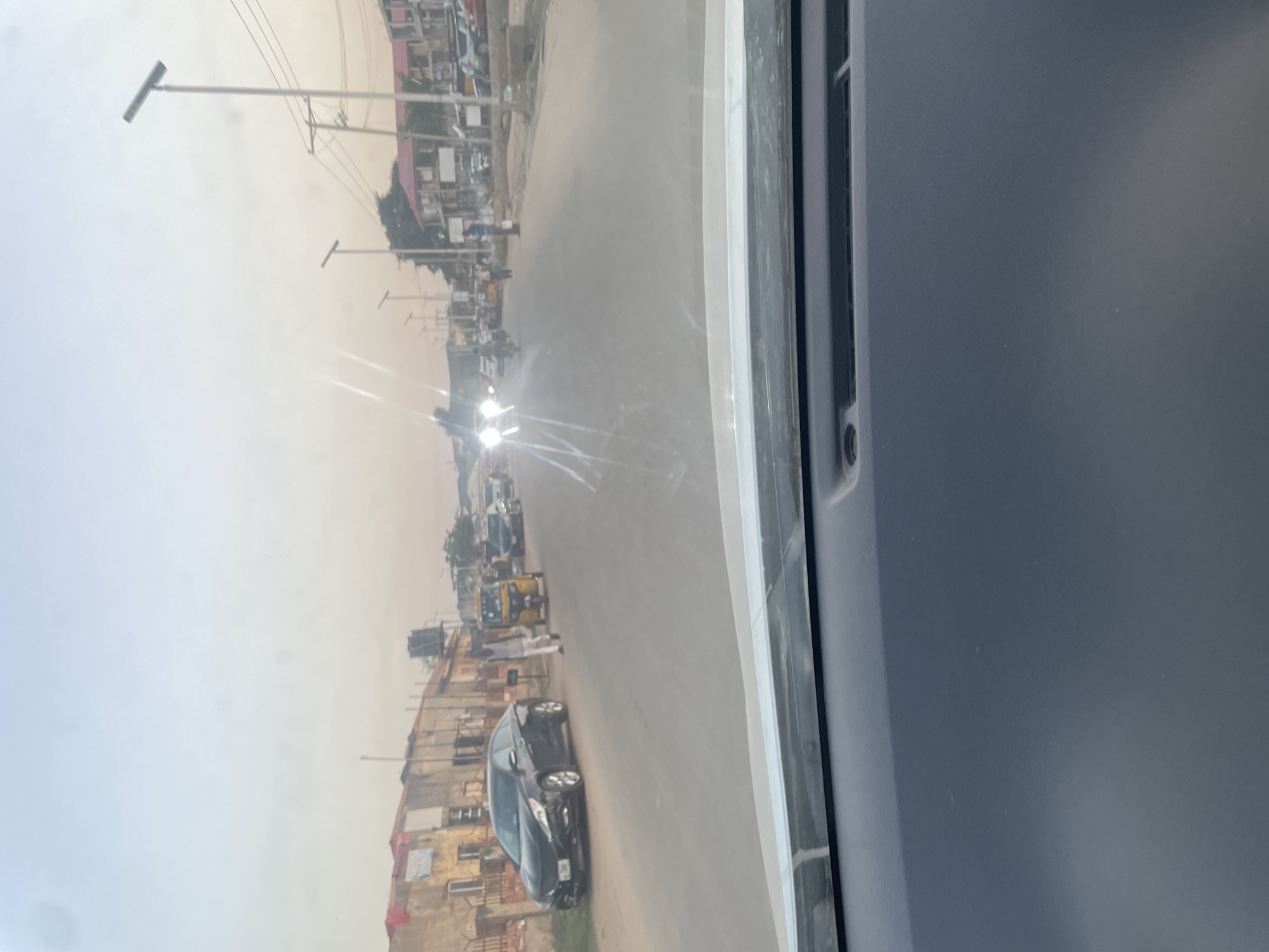 The image depicts a street scene in Abuja, Nigeria, viewed from inside a vehicle. The foreground shows part of the vehicle's interior in gray. The street is filled with vehicles, including a dark-colored car parked on the side of the road, a "keke" (auto-rickshaw), and other unidentified vehicles moving further down the street. People are walking alongside the road. Buildings line the street, appearing to be a mix of residential and commercial structures. Utility poles and power lines are visible above. The sky is overcast and hazy, possibly indicating dusty conditions. The light suggests it is daytime.