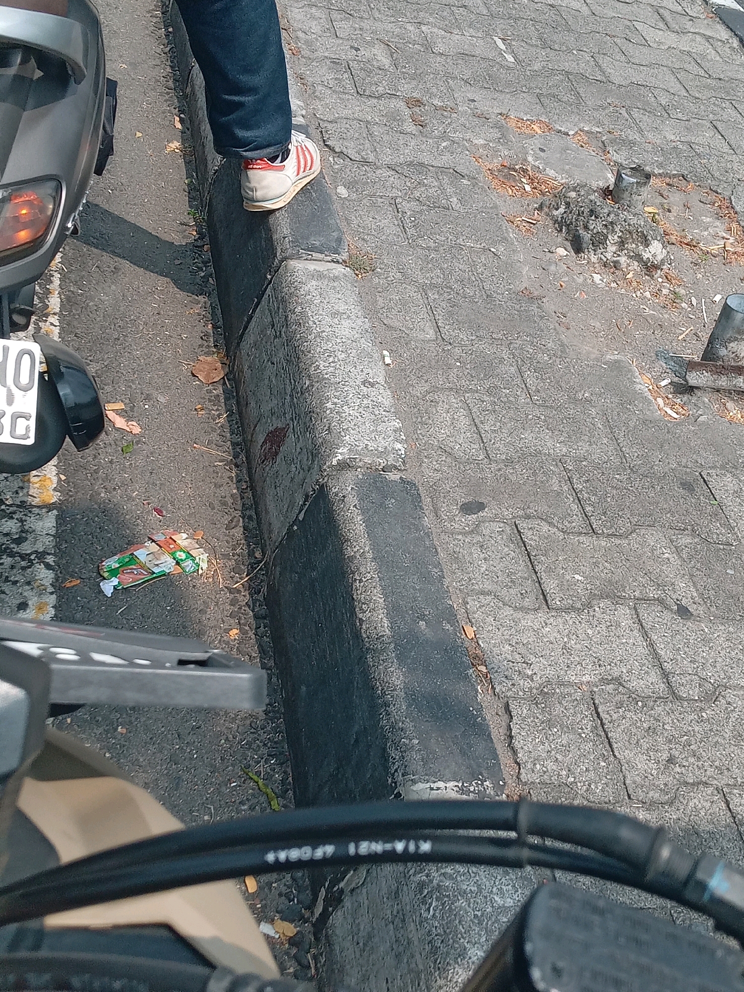 A person is standing on a curb with their right foot wearing a white and red sneaker with the Adidas logo visible. They are wearing dark blue denim jeans. The person is next to a grey scooter which is partially visible. The ground is paved with interlocking grey concrete blocks. There is some litter on the ground, including a crumpled packet of cigarettes. The scene is set on a street in Ngaglik, Indonesia.