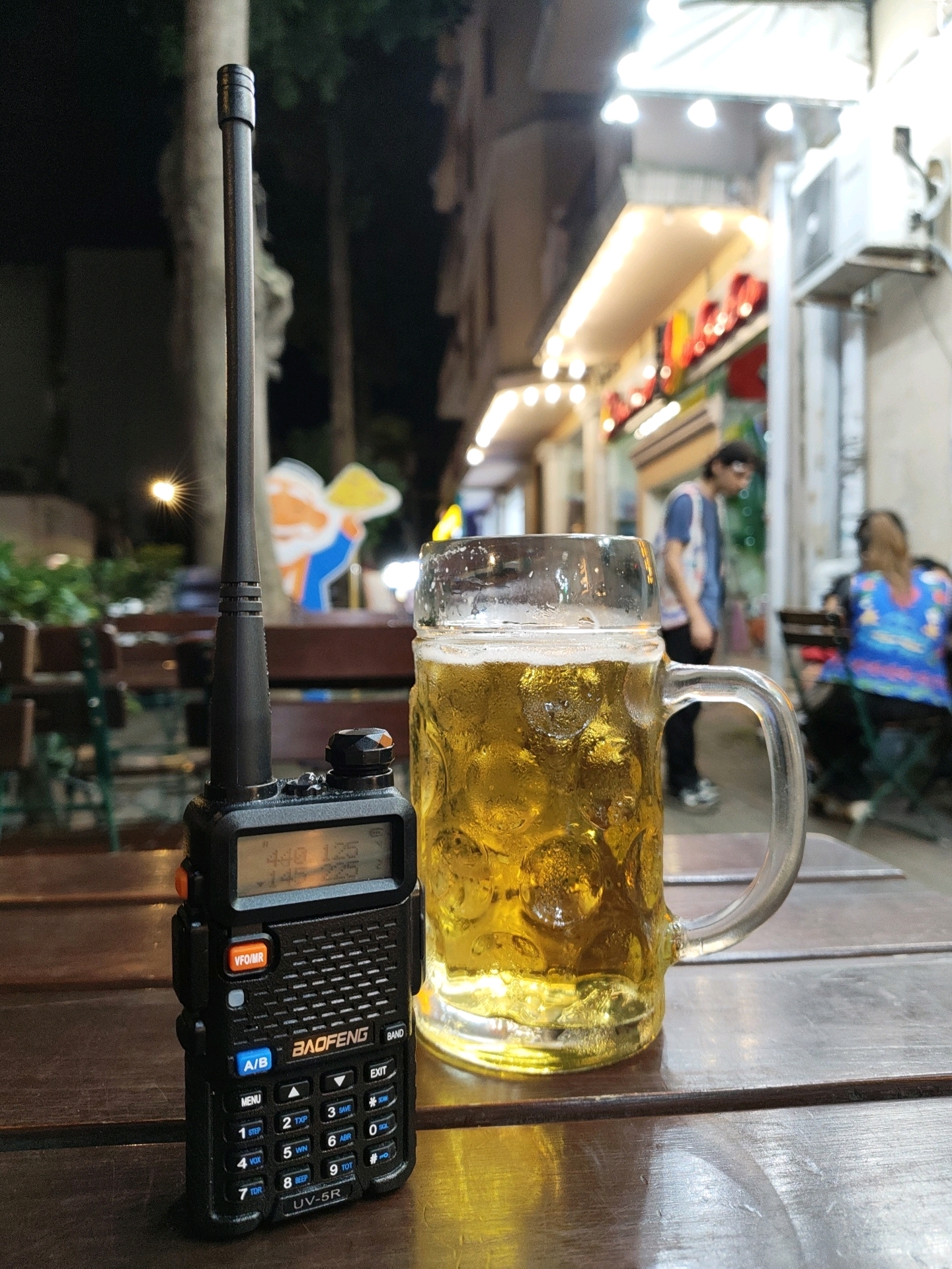 A Baofeng UV-5R walkie-talkie sits on a wooden table next to a tall glass mug filled with a frothy, golden beer. The walkie-talkie's screen displays "446.125". In the background, blurred figures are visible sitting at tables outside what appears to be a bar or restaurant, illuminated by warm, string lights. The scene is set at night on a street in Batumi, Georgia, characterized by its pedestrian-friendly, open-air dining establishments.