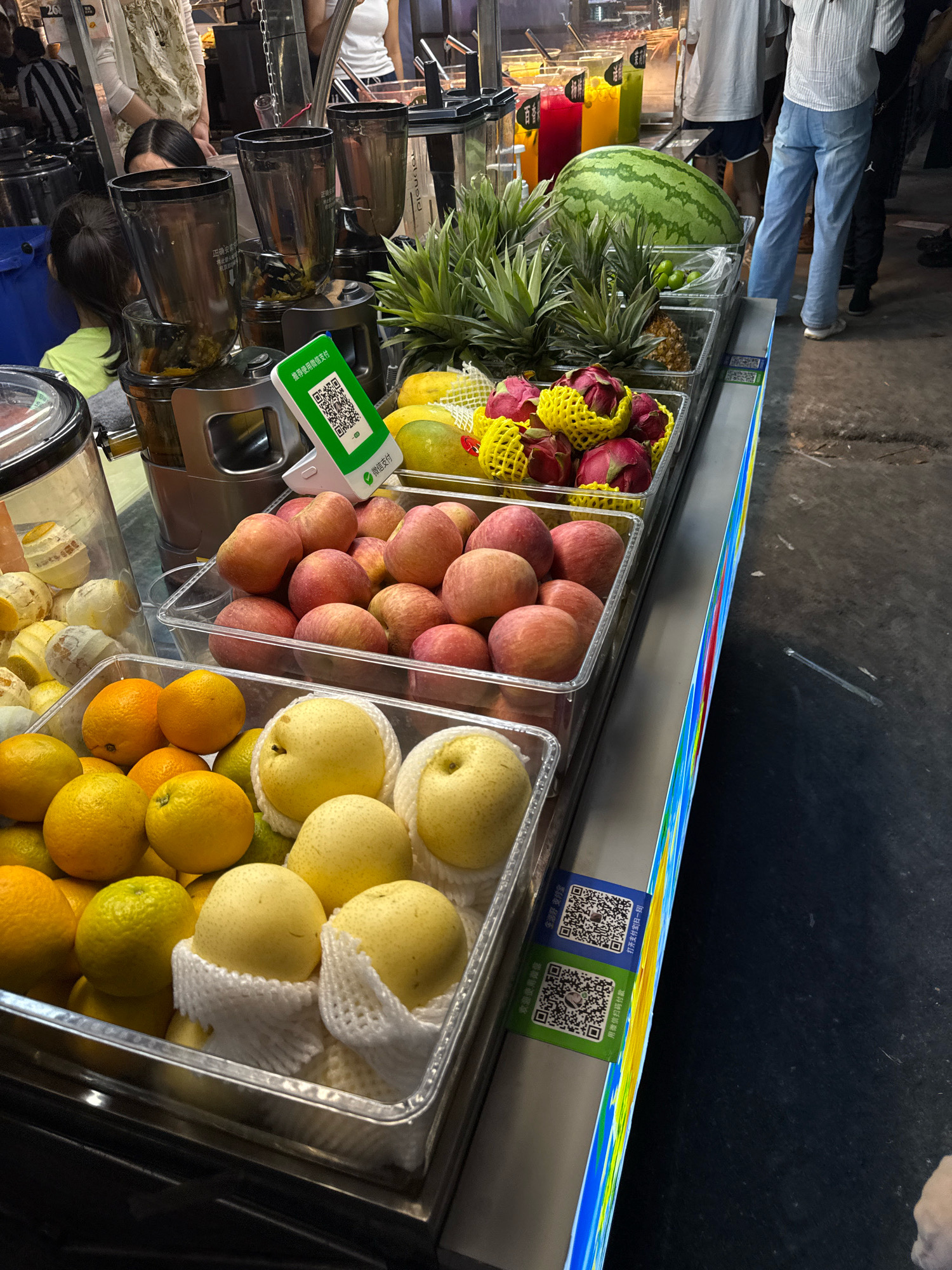 The image depicts a street food stall in Yiwu, China, displaying a variety of fresh fruits for sale. In the foreground, several containers hold assortments of oranges and yellow pears, some of which are wrapped in protective netting. Behind these, a container is filled with red apples. Further back, a display showcases pineapples and dragon fruit, some of which are also netted. A large watermelon is prominently featured. Adjacent to the fruit display are several tall blenders, suggesting that fresh juices are also being prepared and sold. The stall has a colorful, illuminated edge. A small green stand with a QR code is visible on the counter, likely for payment processing. In the background, partially obscured by the stall, are several people, including individuals wearing casual clothing and one person in a white shirt and blue jeans. The overall scene suggests a bustling market environment.