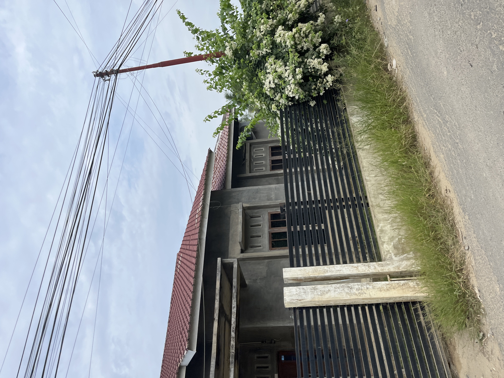 The image shows a house in Tanahterban, Indonesia. The house has a red tiled roof and gray concrete walls. It is surrounded by a black metal fence. In front of the fence, there is a large bush with white flowers. The sky is cloudy. A red utility pole with many wires runs across the top of the image. The road in front of the house is paved. There are no people visible in the image.