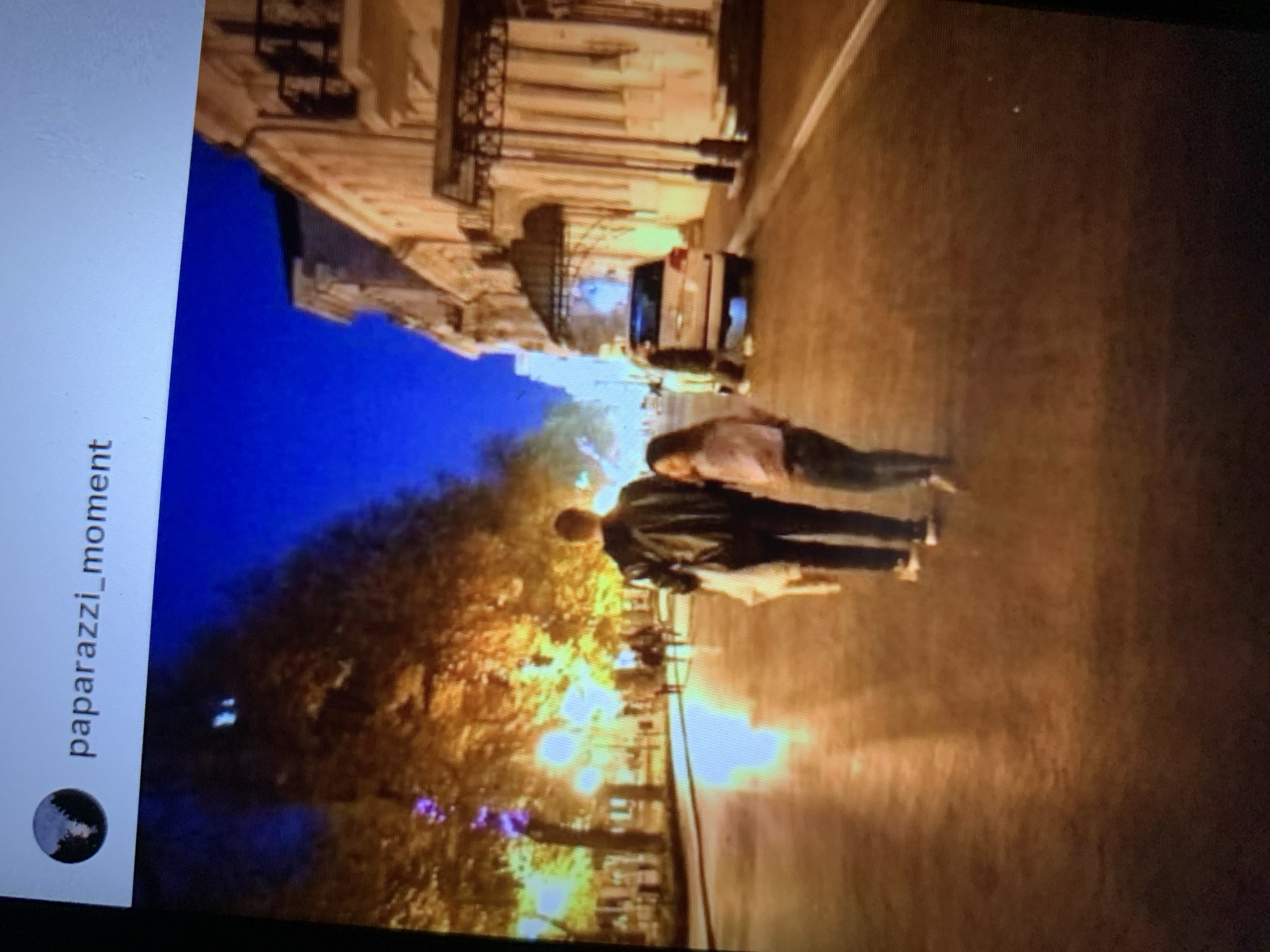 The image is a screen capture of a social media post with the username "paparazzi_moment" visible in the top left corner. The scene depicts a nighttime street in Lymanka, Ukraine, illuminated by streetlights and the glow of buildings. A man in a black jacket and dark pants is walking away from the camera, carrying a white bag. Beside him walks a young woman in a light-colored top and dark pants, also facing away from the camera. They are walking down a wet street, reflecting the lights, with trees lining one side of the street and buildings on the other. A white SUV is parked further down the street. The atmosphere is calm and somewhat romantic due to the nighttime setting and soft lighting.