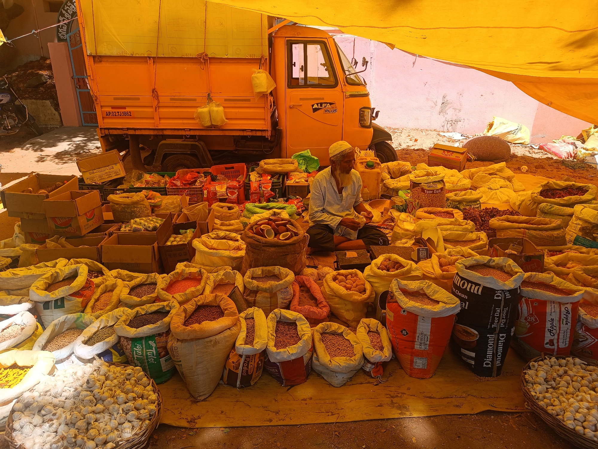 Here's a detailed description of the image:
**Location and Environment:**
The image appears to be taken at an outdoor market or trading location in Enumulapalli, India, with many different kinds of bags of ingredients, spices, and garlic. A yellow tarpaulin provides shade. The ground is a rough surface, possibly earth or pavement.
**People:**
An older man with a white beard and cap is seated in the middle of the frame. He appears to be focused on a task, possibly sorting or preparing items for sale.
**Subjects and Objects:**
* The foreground is dominated by a variety of bags filled with grains, lentils, beans, spices, and vegetables, displayed for sale. There are various sizes and colors of bags.
* Cardboard boxes containing items.
* An orange three-wheeled truck (an "Alfa Plus") is visible in the background, partially obscured by the tarpaulin. It is likely used for transporting goods.
* A few coconuts are also visible.
**Actions and Interactions:**
The primary action is the man's activity of sorting or preparing goods. Customers are likely intended to purchase the items on display. The truck suggests the transport and supply of the market items.