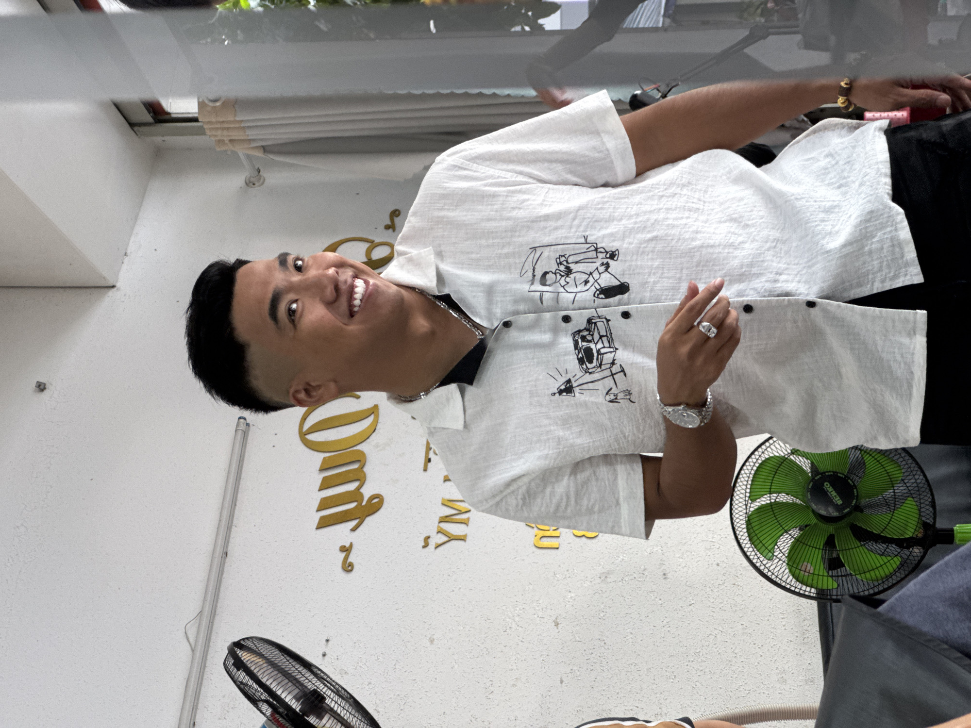 A young Asian man with a fade haircut and a bright smile poses for a photo in what appears to be a barbershop or salon in Nha Trang Ward, Vietnam. He is wearing a white short-sleeved button-up shirt with a black graphic print on the front, a silver chain necklace, a silver watch on his left wrist, and a large silver ring on his left index finger. The background features a white wall with decorative gold lettering and two fans, one black with a silver cage and the other green and black. A window with sheer curtains is visible to the right of the man, reflecting the outdoor environment. The man is looking towards the camera and his body is angled slightly to the left.
