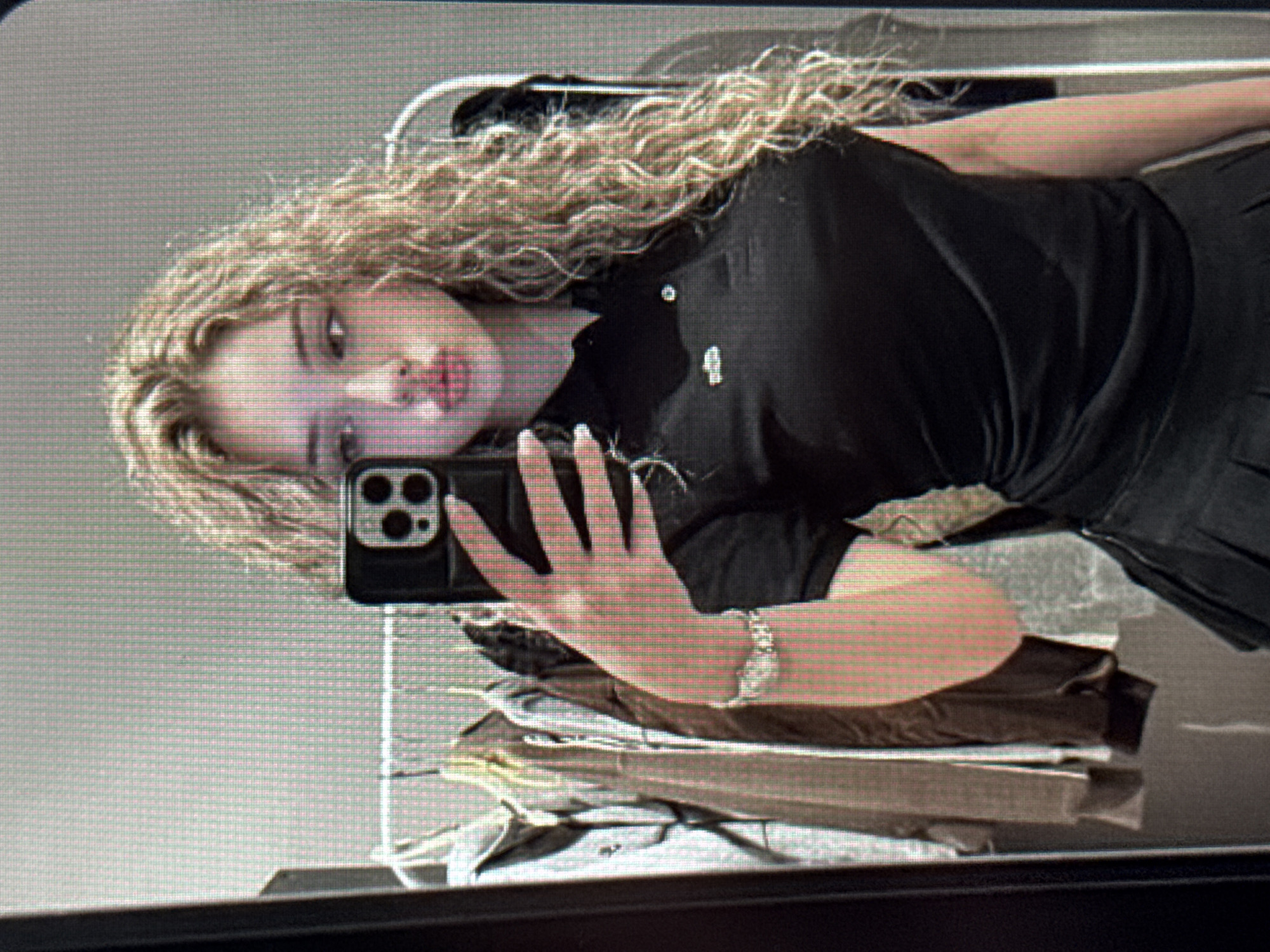 A young woman with long, voluminous, curly blonde hair is captured in a mirror selfie, with the image rotated 90 degrees counter-clockwise. Her head is positioned on the left side of the frame, tilted slightly upward, with her body extending to the right. She is wearing a black polo shirt with a visible white logo or emblem on the chest and a dark-colored skirt or bottom. A metallic watch is worn on her left wrist.
The woman is holding a dark-colored smartphone, possibly an iPhone due to the distinct camera module design, in her left hand, pointing it towards the mirror to capture her reflection. Her face is visible, featuring full lips slightly parted, and her gaze is directed towards the camera. The background, also a reflection, reveals an indoor setting. Behind her, a white or light-colored clothing rack holds garments, and beneath her arm, a stack of folded items, possibly clothes or fabric, is visible on a surface. The overall image has a somewhat pixelated or low-resolution quality, characteristic of a digital screen capture. There are no discernible location cues within the image to specify "Varna, Bulgaria." No legible text is visible in the frame.