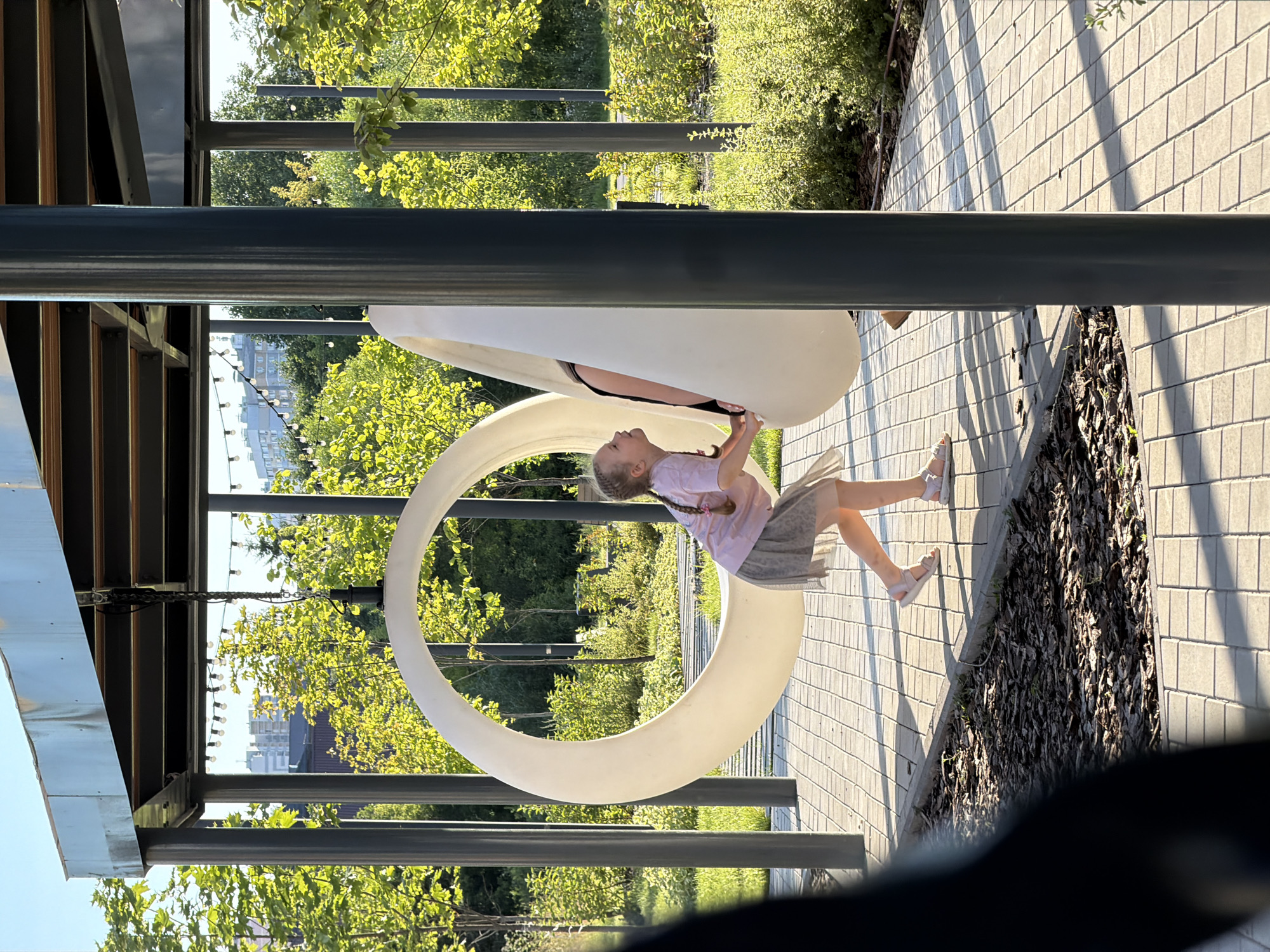 In this media file, a young girl with braided blonde hair, wearing a light purple t-shirt and a grey skirt, is on a playground. She is positioned inside a large, circular, white structure, possibly a swing or play element, and appears to be in mid-air, suggesting she is swinging. Her left hand is reaching up towards the top of the circular structure, and her right hand is holding onto the inside edge. She is looking to her right with a slight smile, indicating a joyful or excited emotion. Her legs are bent, with one foot slightly in front of the other. The playground has a paved ground made of light grey bricks, casting long shadows from the surrounding structures and trees, suggesting it is late afternoon or early evening. Lush green trees and bushes are visible in the background, interspersed with some buildings in the distance. The setting appears to be an outdoor public space, likely a park or recreational area in Kazan, Russia, given the provided location context. The overall atmosphere is bright and sunny, contributing to a pleasant and cheerful mood. There is no visible text in the image.