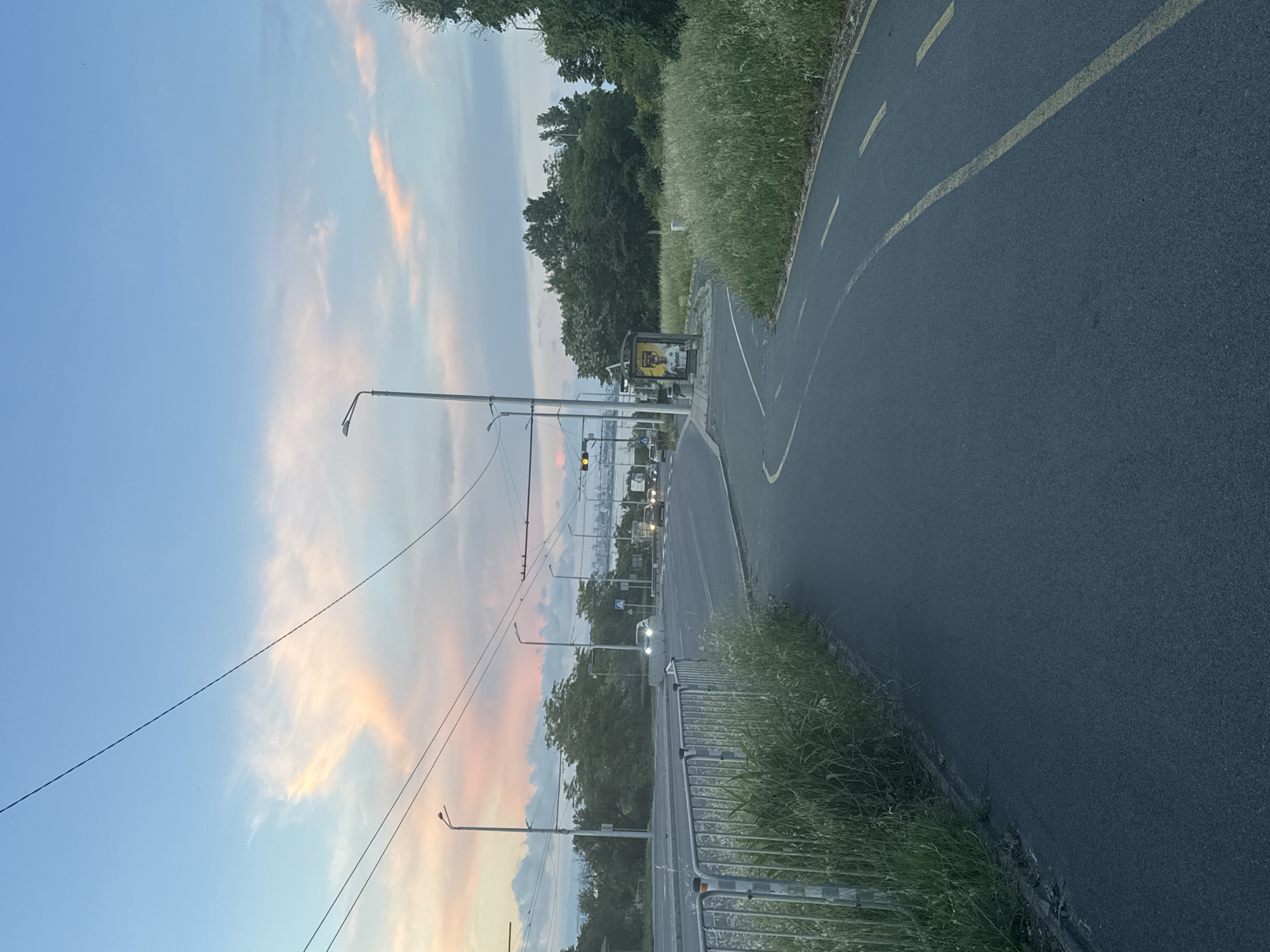 The image depicts a paved bicycle path on the left side of a road in Burgas, Bulgaria, at dusk. The sky is a clear blue with wisps of pink and orange clouds. A few trees and streetlights line the road, which has several cars driving on it. In the distance, a body of water can be seen, possibly the Burgas Bay. A bus stop with an advertisement is also visible on the right side of the road. The bicycle path has yellow lines marking its lanes. The overall impression is of a serene urban landscape at the end of the day.