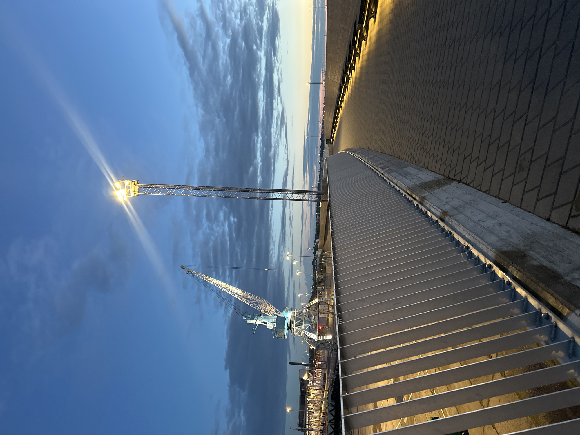 This image captures a twilight scene along a waterfront promenade in Tallinn, Estonia. A large, illuminated industrial crane stands prominently on the left side of the frame, its white and blue structure contrasting with the darkening sky. To the right, a tall, metal tower with bright lights illuminates the pathway, which is paved with dark cobblestones laid in a diamond pattern. The promenade itself is bordered by a modern, light-colored metal railing and a concrete retaining wall. In the distance, a cityscape with more lights and structures is visible, suggesting an urban port environment. The sky is a deep blue, with scattered clouds catching the last rays of the setting sun, creating a soft glow on the horizon. The overall impression is one of industrial activity and public infrastructure at dusk.