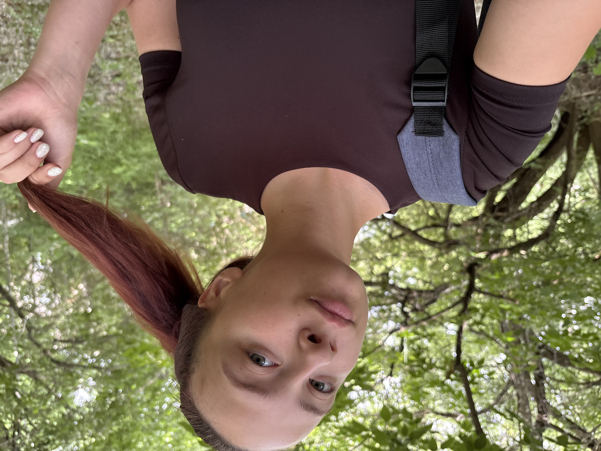 The image is a vertical, medium shot of a young woman outdoors, with her head tilted downwards as if looking up at the camera. She has fair skin, green eyes, and light brown hair with reddish undertones, pulled back into a ponytail. She is wearing a dark brown t-shirt and appears to be holding a strand of her hair. The background is filled with lush green foliage, suggesting a forest or wooded area. The lighting indicates it's daytime, possibly late morning or afternoon, with soft light filtering through the leaves. The overall impression is casual and relaxed. There is no visible text. The setting, with its dense greenery and tree branches, suggests a natural outdoor environment. The location context is Chaikovsky, Russia.