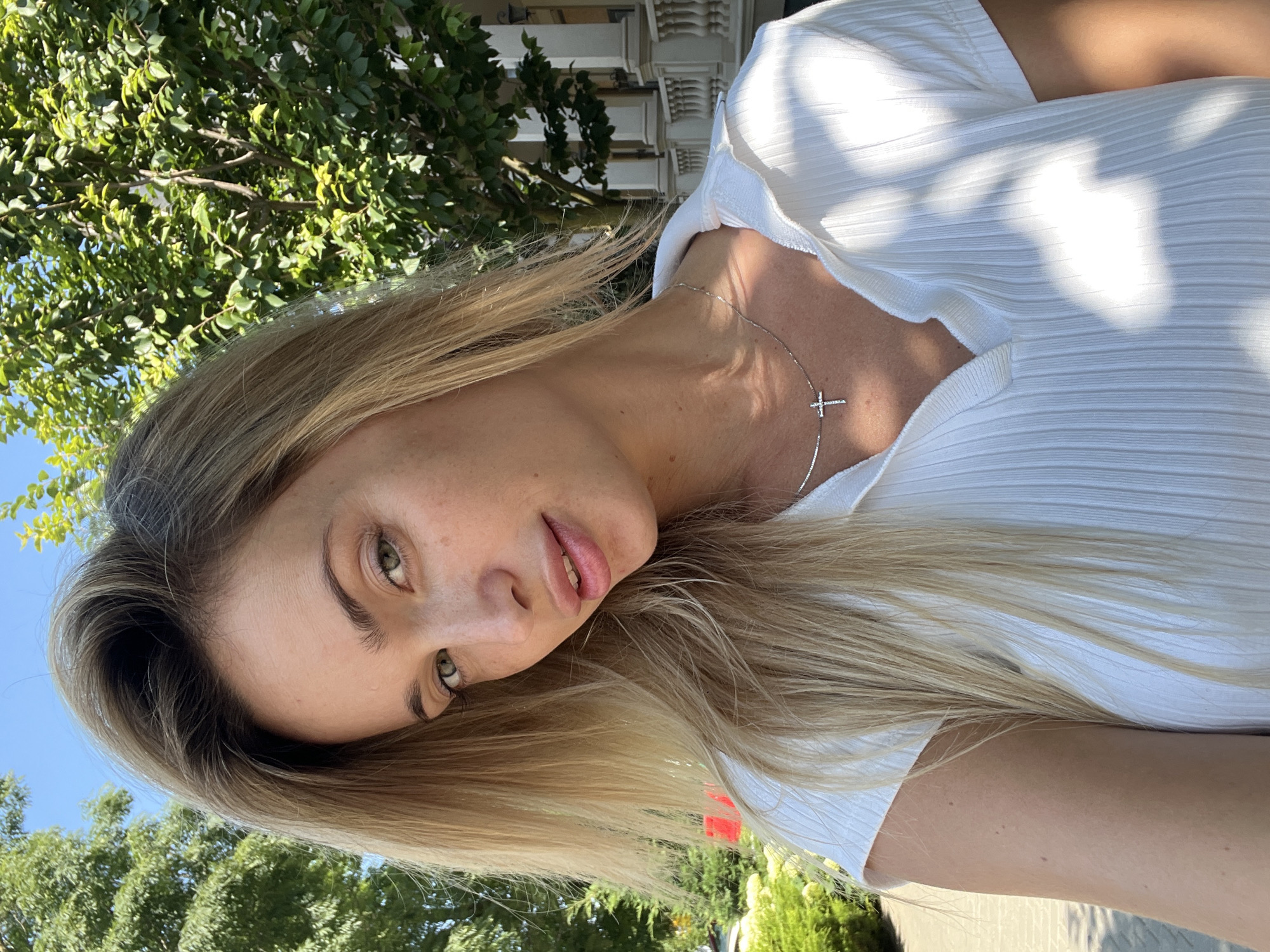 A blonde woman with light green eyes poses for a selfie outdoors. She is wearing a white, ribbed shirt with a collar and a silver necklace with a cross pendant. Her hair is long and falls over her shoulders. The background includes lush green trees, a bright blue sky, and architectural elements of a building, suggesting an urban or suburban setting. The lighting indicates it is daytime.