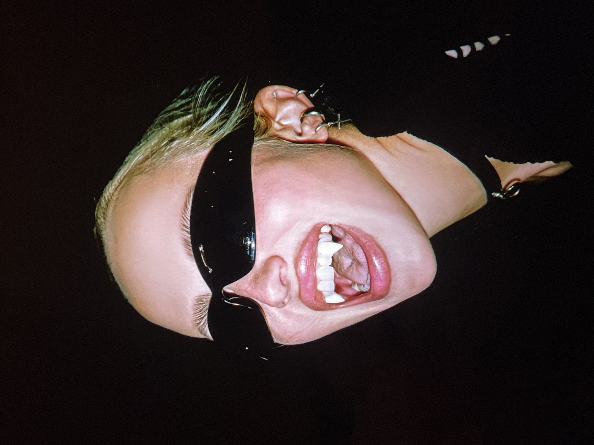 This is a close-up, upside-down, studio portrait of a person against a black background. The subject has blonde hair, and their face is partially obscured by a black, shiny material, possibly latex or paint, covering their left eye and cheek. The visible right side of their face shows an open mouth with bright pink lips and white teeth, some of which appear to be dental implants or caps. The person has earrings in their right ear, including a hoop and a cross-shaped charm. The lighting is dramatic, highlighting the textures of the skin and the glossiness of the black material. The overall mood is intense and somewhat unsettling, with the subject's open mouth suggesting a scream or gasp. There is no discernible text in the image. The context of Saint Petersburg, Russia, does not provide specific visual cues for this abstract portrait.