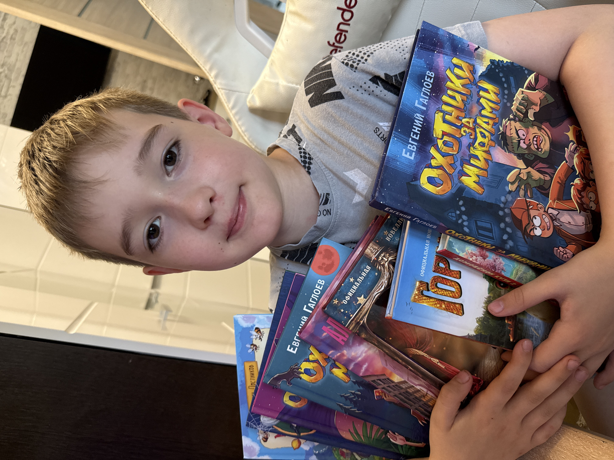 A young boy with light brown hair and brown eyes is holding a stack of Russian books. He is wearing a grey t-shirt with a black graphic print. The boy is seated in a white chair, possibly a gaming chair, with the word "Defenders" visible on the headrest. The background appears to be indoors, with a dark, possibly wooden surface on the left and a light-colored wall with reflective panels to the right. The books have colorful illustrations and Russian text on their covers. One visible book title is "Охотники за мифами" (Hunters for Myths). The image is taken from a slightly low angle, focusing on the boy and the books he is holding. The setting does not provide specific details to pinpoint a location within the city of Oktyabrskoye, Russia.