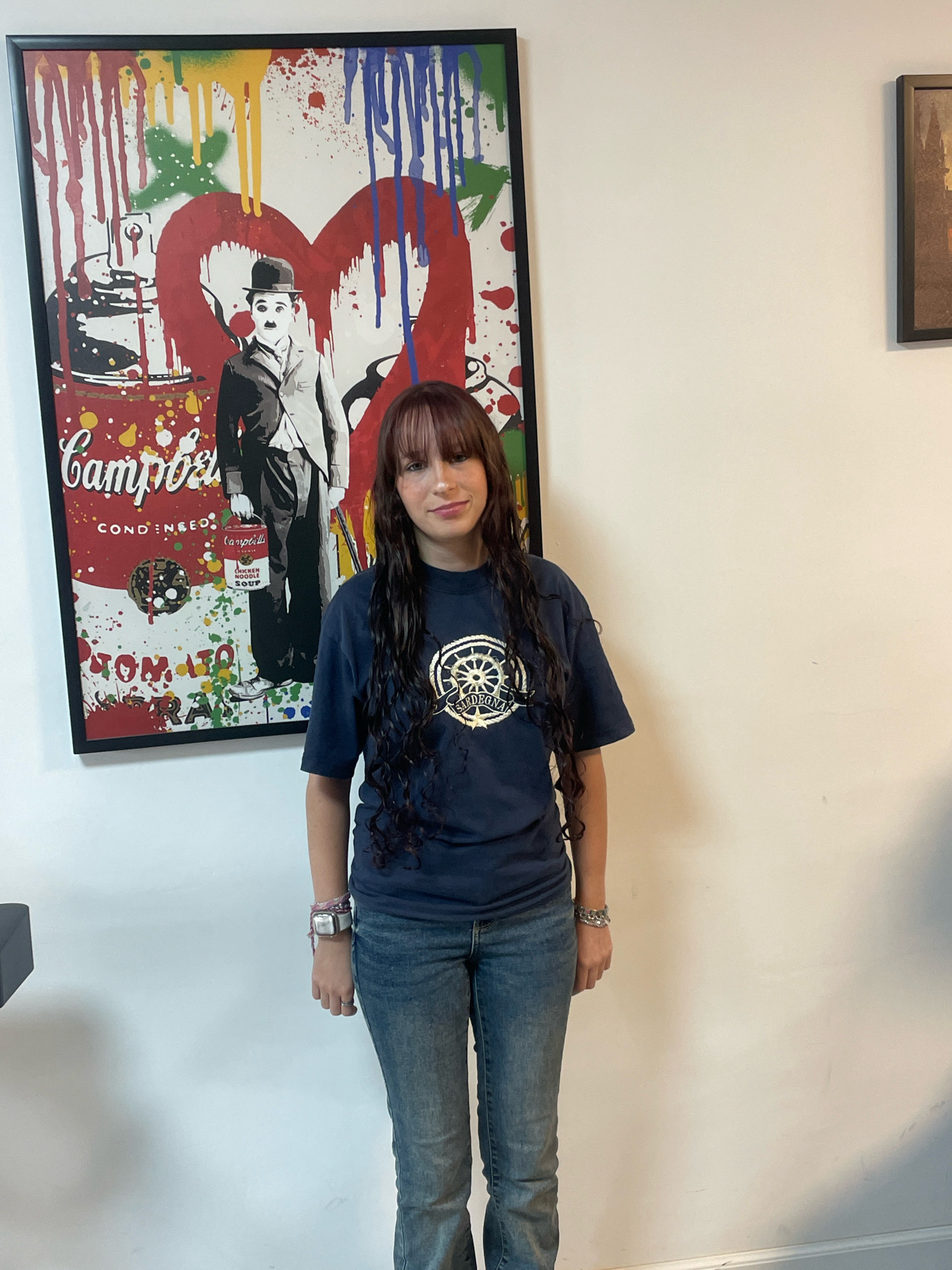 A young woman stands in front of a large framed artwork featuring a colorful, abstract depiction of a Campbell's soup can and Charlie Chaplin. She has long, dark, wavy hair and is wearing a dark blue t-shirt with a circular emblem on it, and blue jeans. She is also wearing a watch on her left wrist and a bracelet on her right wrist. The artwork is on a white wall, and a dark framed picture is visible on the wall to the right. The scene appears to be indoors.